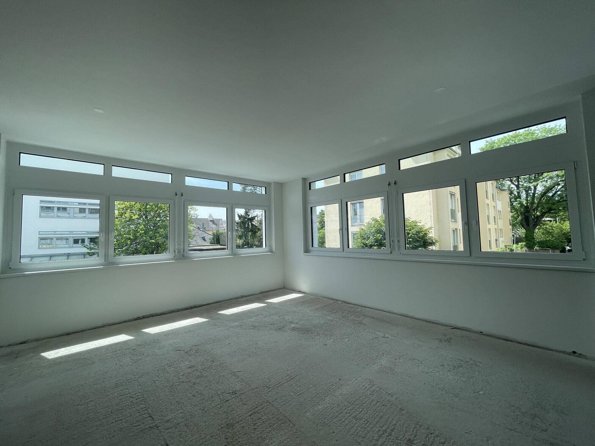 Heller Büroraum mit durchgehenden Fenstern und Blick auf umliegende Gebäude und Bäume, sehr lichtdurchflutet.