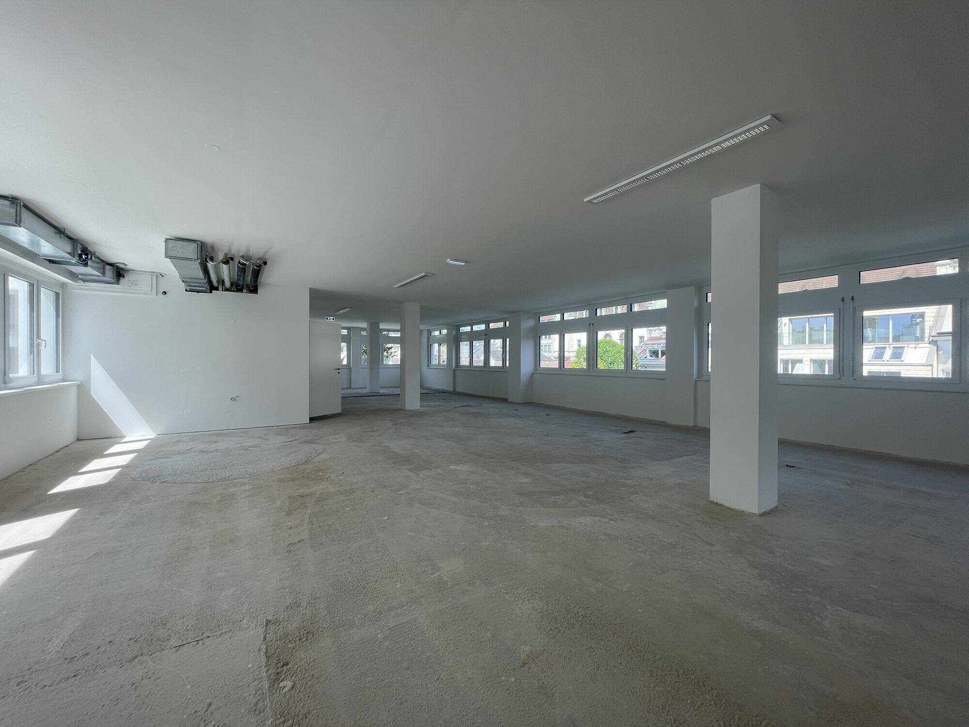 Großer, heller Büroraum mit sichtbaren Lüftungsrohren und vielen Fenstern.