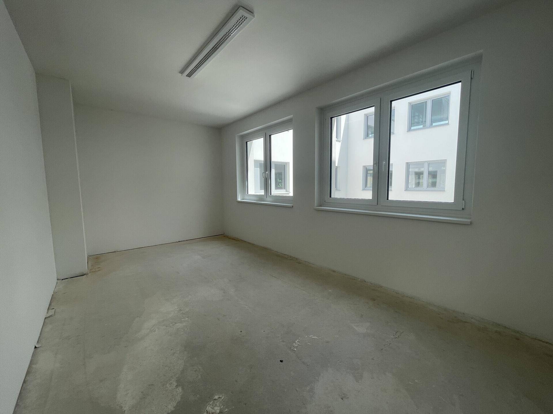 Kleinerer, heller Büroraum mit zwei Fenstern und Betonboden, ideal für ein Einzelbüro.