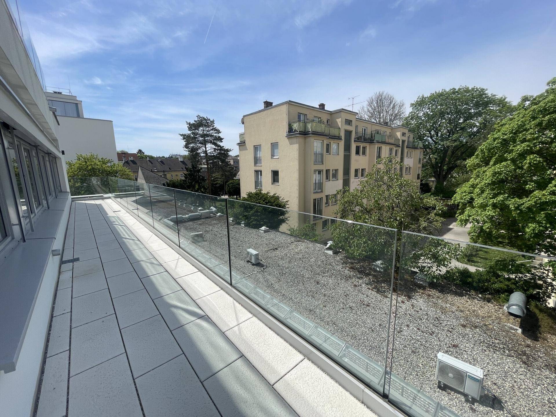 Große Dachterrasse mit Glasgeländer und Blick auf umliegende Gebäude und Bäume.