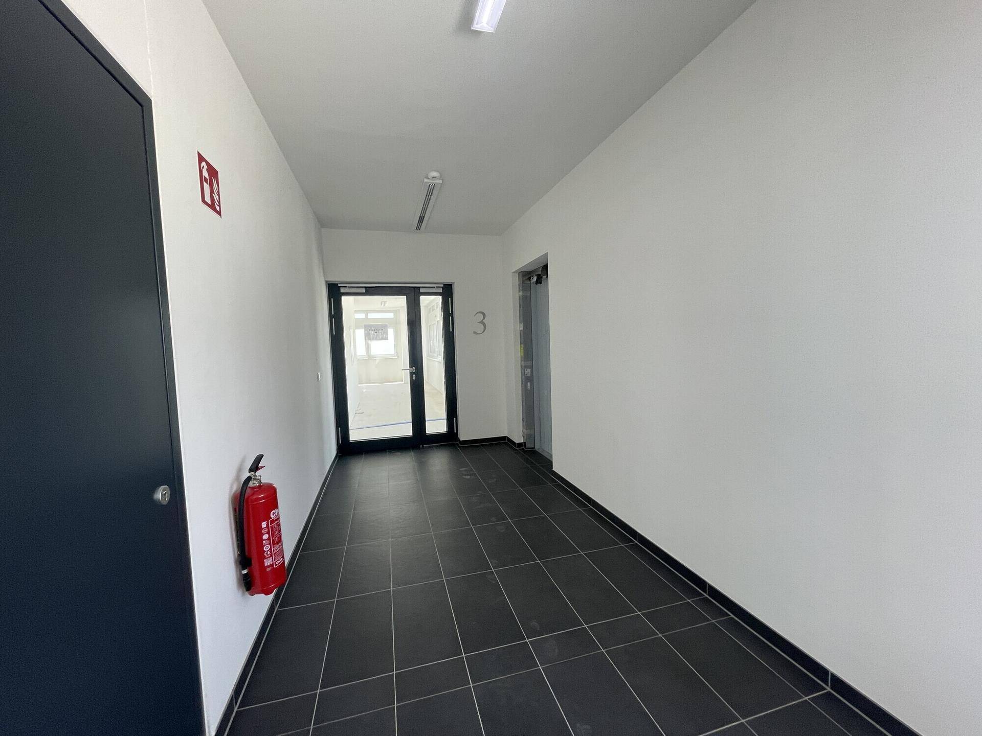 Ein sauberer Flur mit dunklen Fliesen, weiß gestrichenen Wänden und einem Feuerlöscher an der Wand.