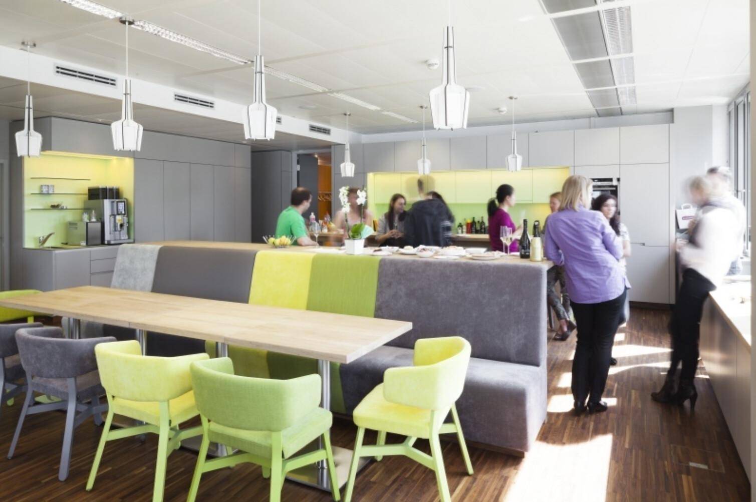 Große, moderne Büroküche mit vielen Personen, langer Theke und hellgrünen Stühlen.