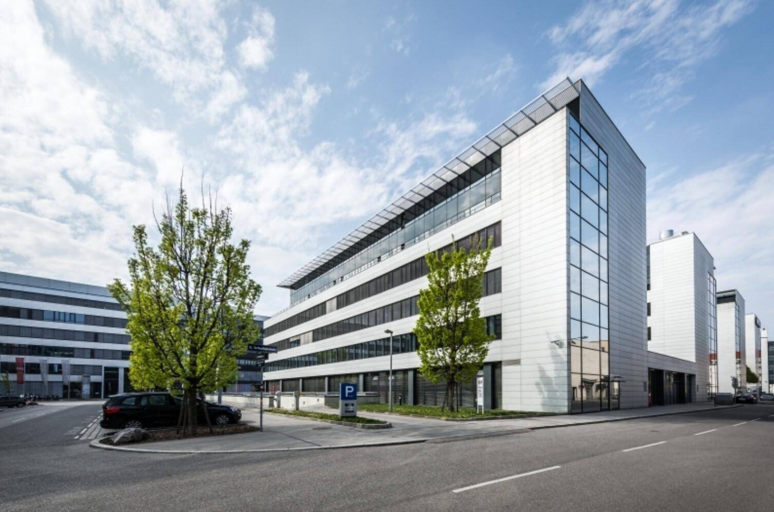 Modernes Bürogebäude mit heller Fassade, großen Fenstern und Bäumen entlang der Straße.