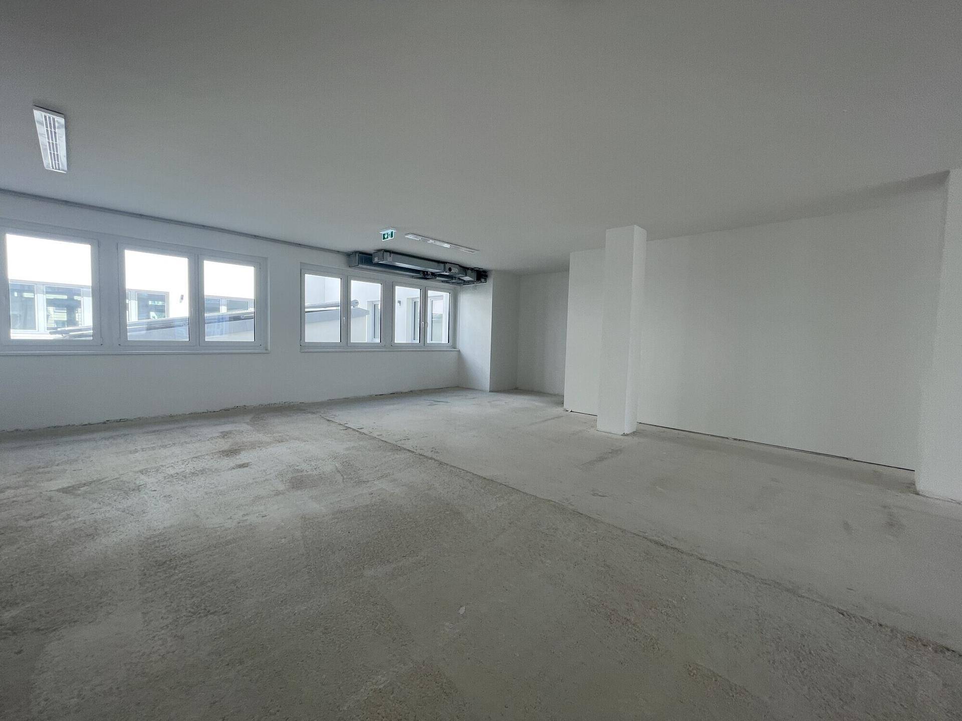 Leerer Büroraum mit großen Fenstern und sichtbarer Lüftungsanlage an der Decke, bereit für die Einrichtung.