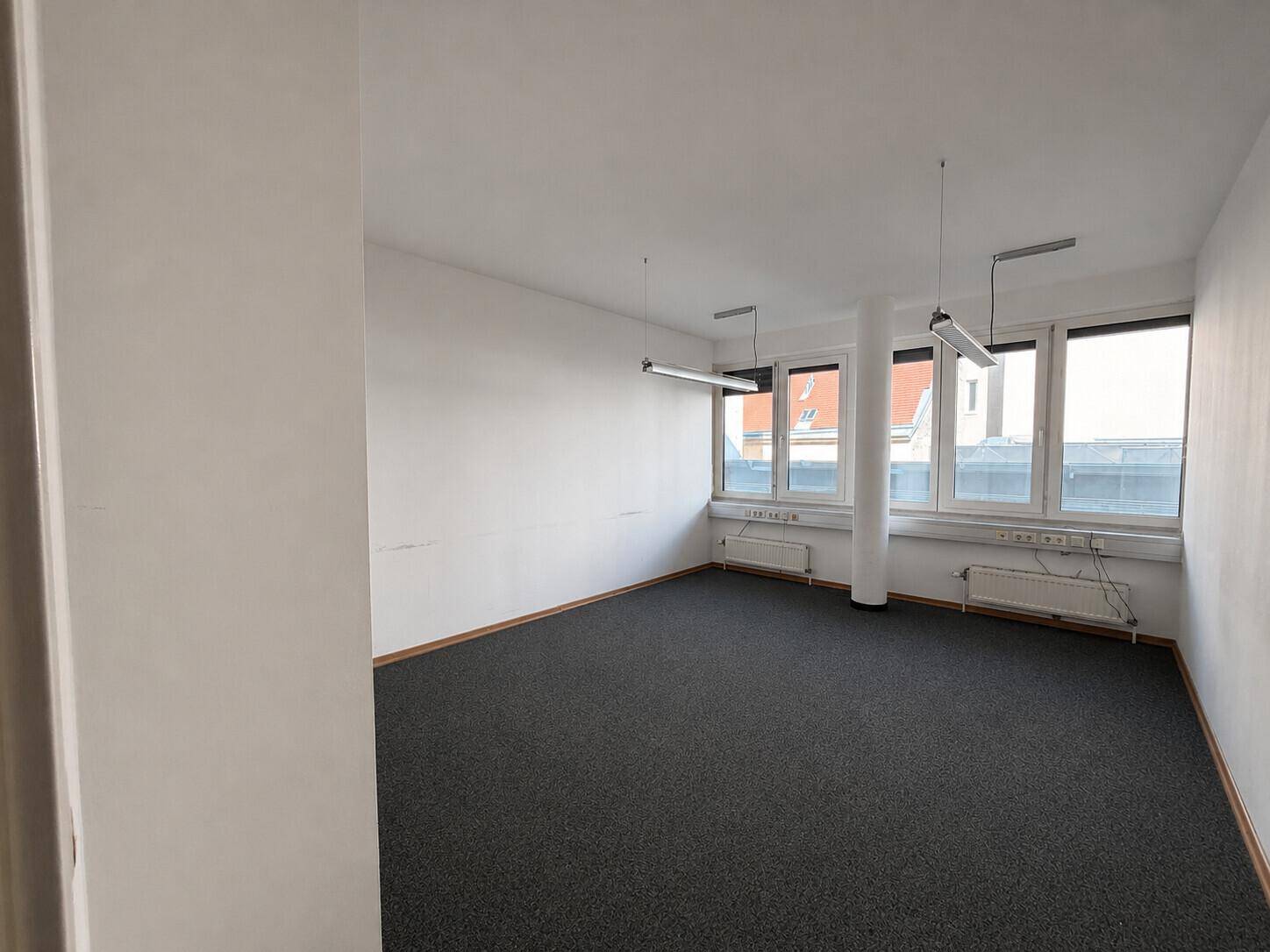 Büro mit Teppichboden, weißen Wänden und großen Fenstern, die den Raum gut beleuchten.