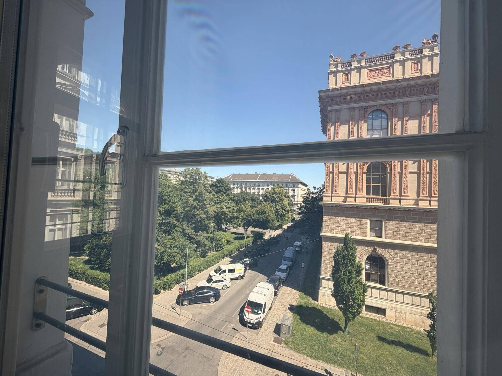Blick aus einem Fenster auf eine belebte Straße mit Bäumen und historischen Gebäuden unter blauem Himmel.