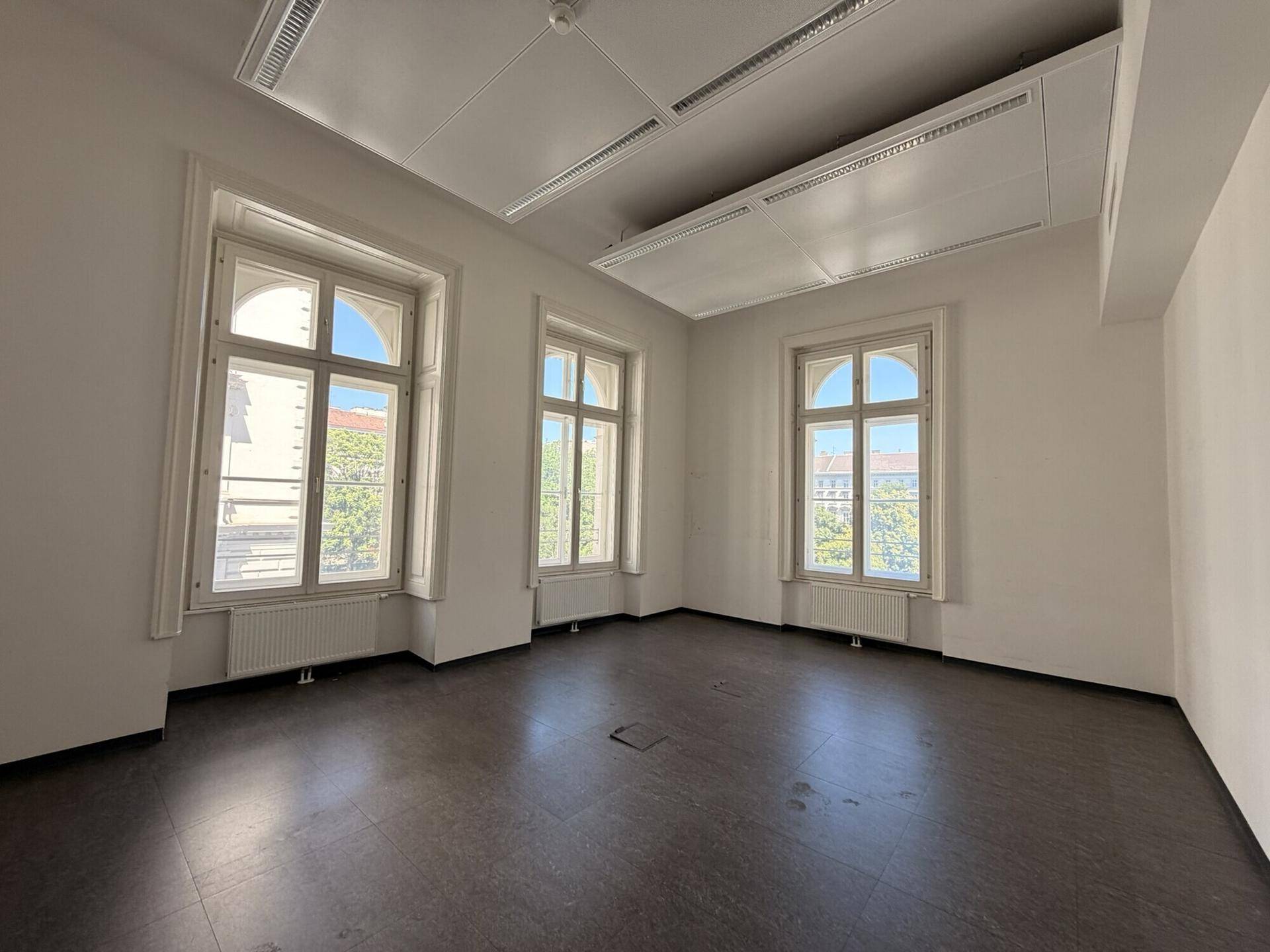 Helles, leeres Büro mit dunklem Bodenbelag, weißen Wänden und drei hohen Bogenfenstern.