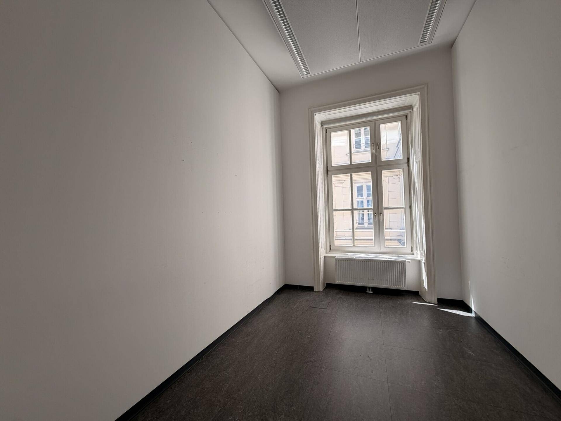 Kleiner, heller Raum mit dunklem Bodenbelag, weißen Wänden und einem hohen Bogenfenster.