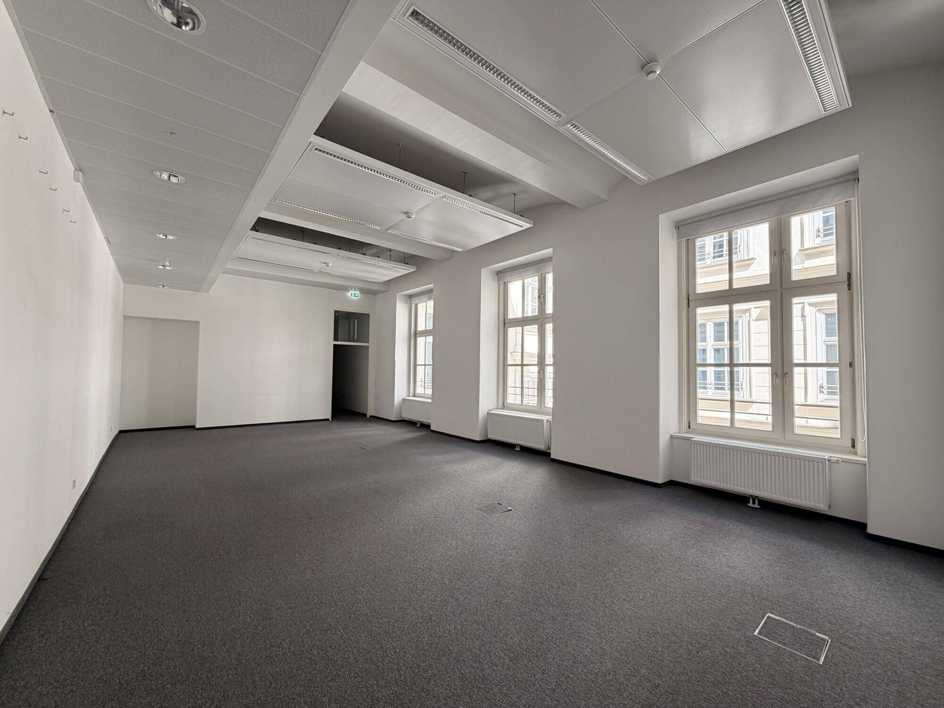 Geräumiges, leeres Büro mit dunklem Teppichboden, vielen Bogenfenstern und einer abgehängten Decke mit Beleuchtung.