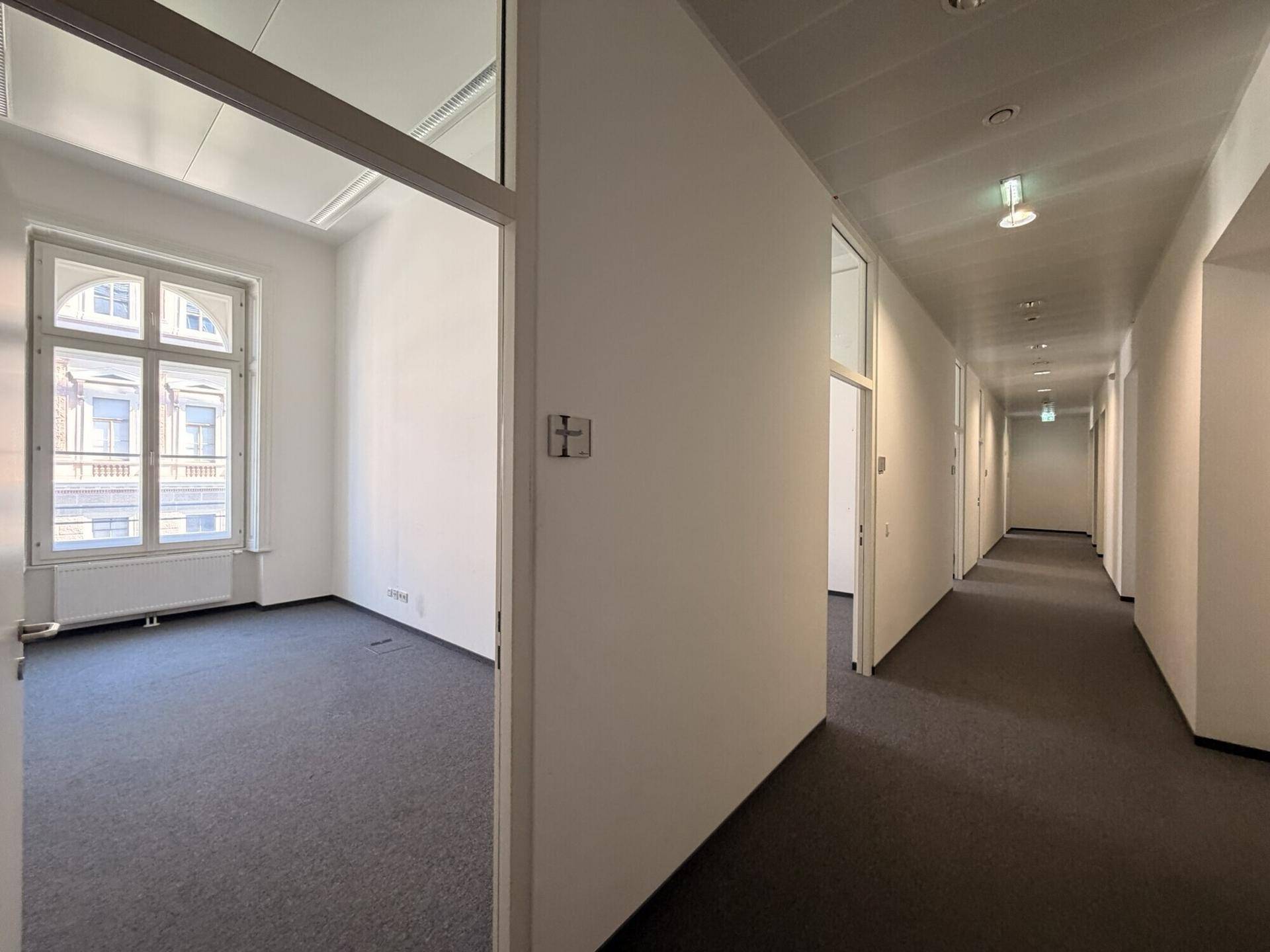 Langer Flur mit Teppichboden und abgehängter Decke, der in ein helles Büro mit Fenster führt.