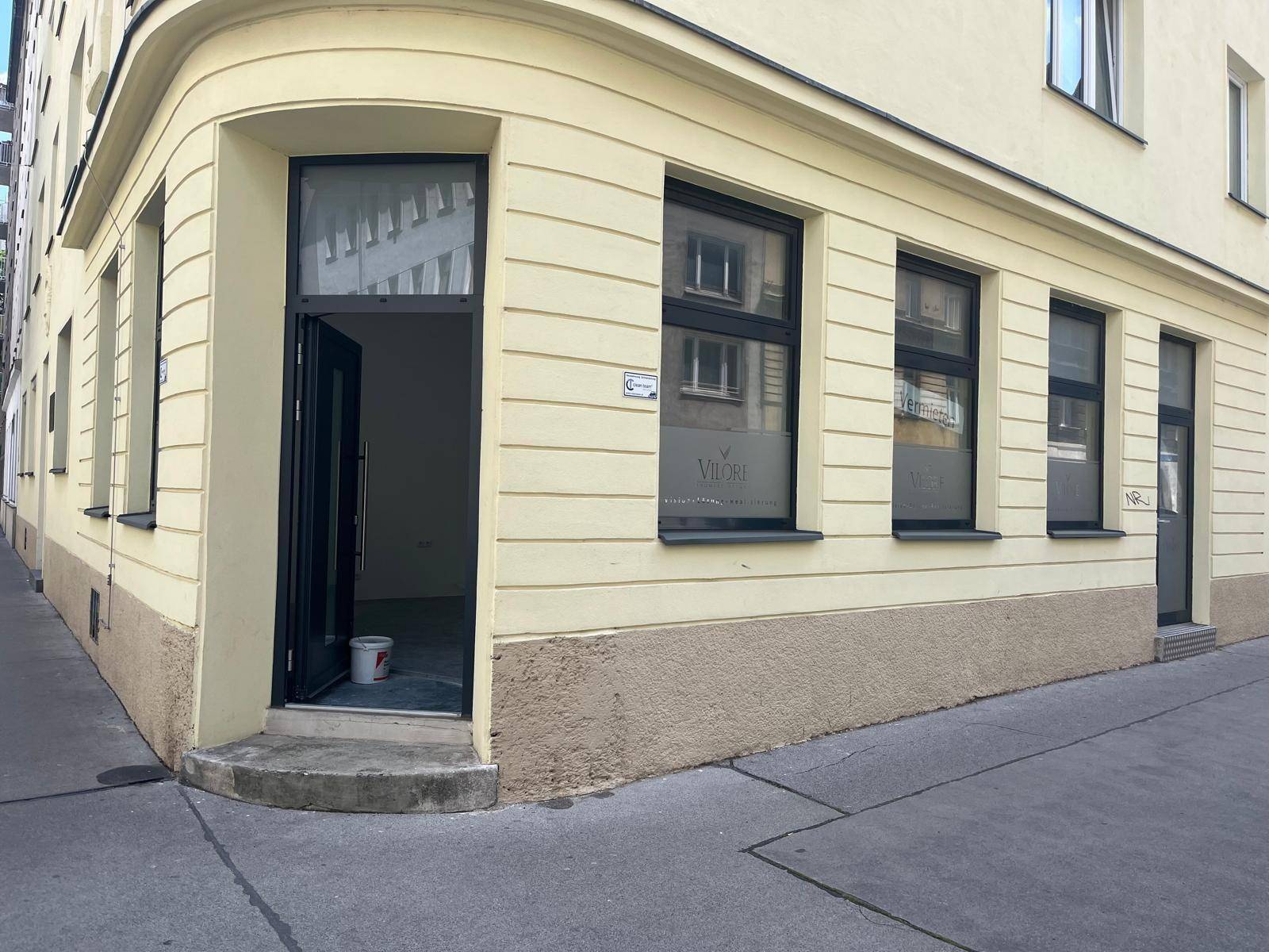 Eckgebäude mit hellgelber Fassade, großen Fenstern und offenem Eingangsbereich auf Straßenniveau.