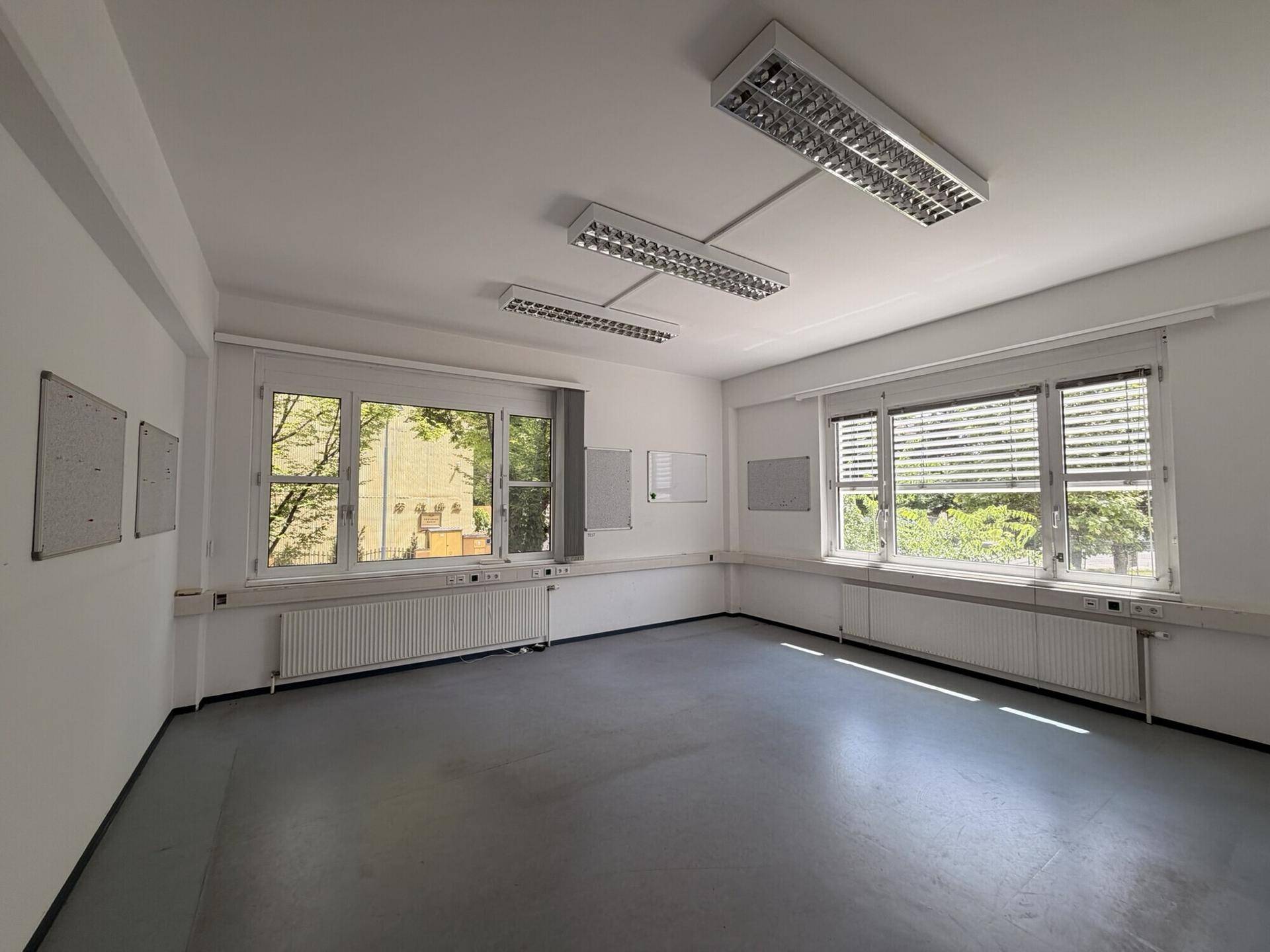 Helles, leeres Büro mit großen Fenstern, die viel Tageslicht hereinlassen und einen grünen Ausblick bieten.