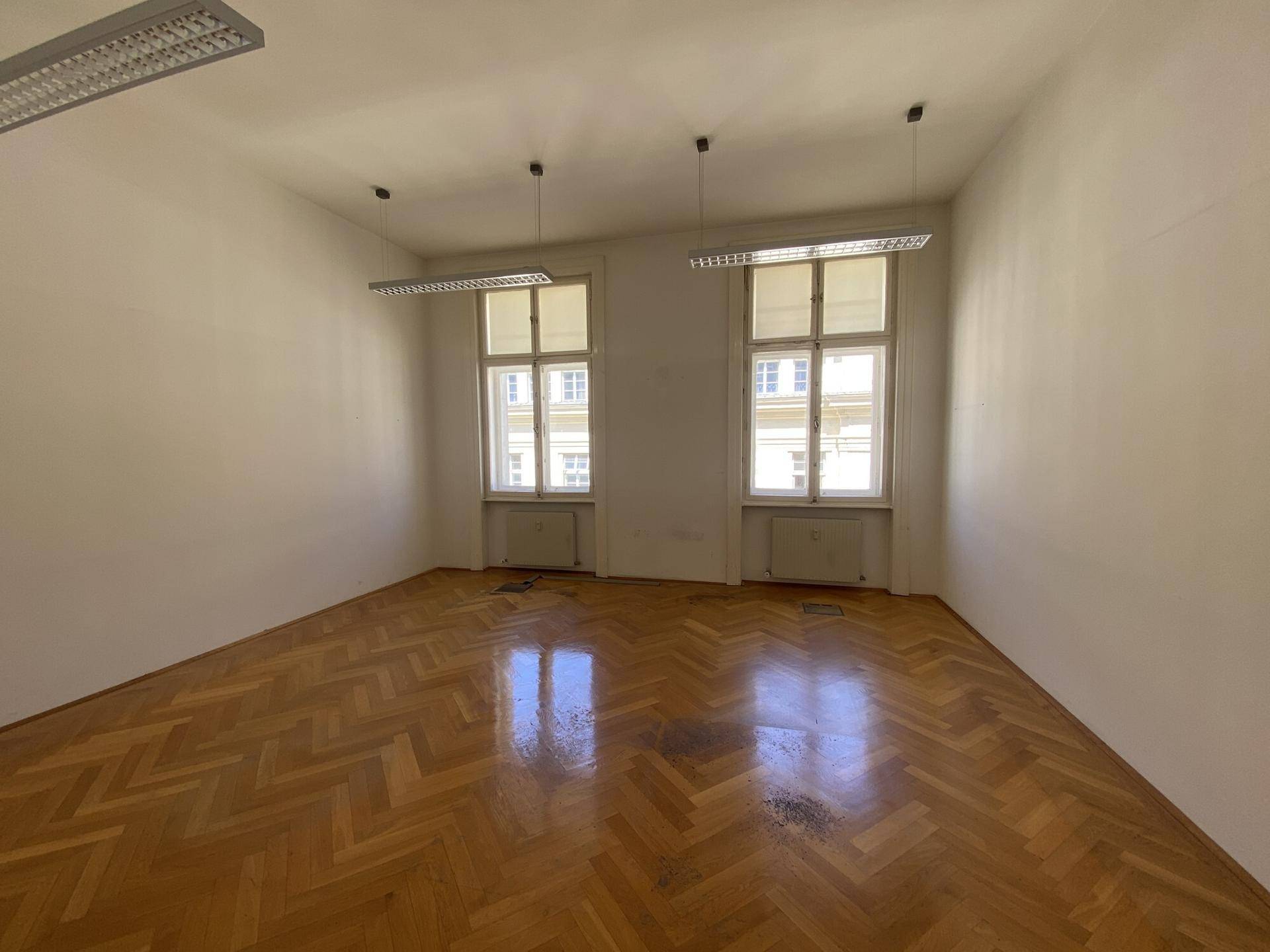 Geräumiges Büro mit glänzendem Fischgrätparkett und zwei hohen Fenstern, die viel Licht hereinlassen.