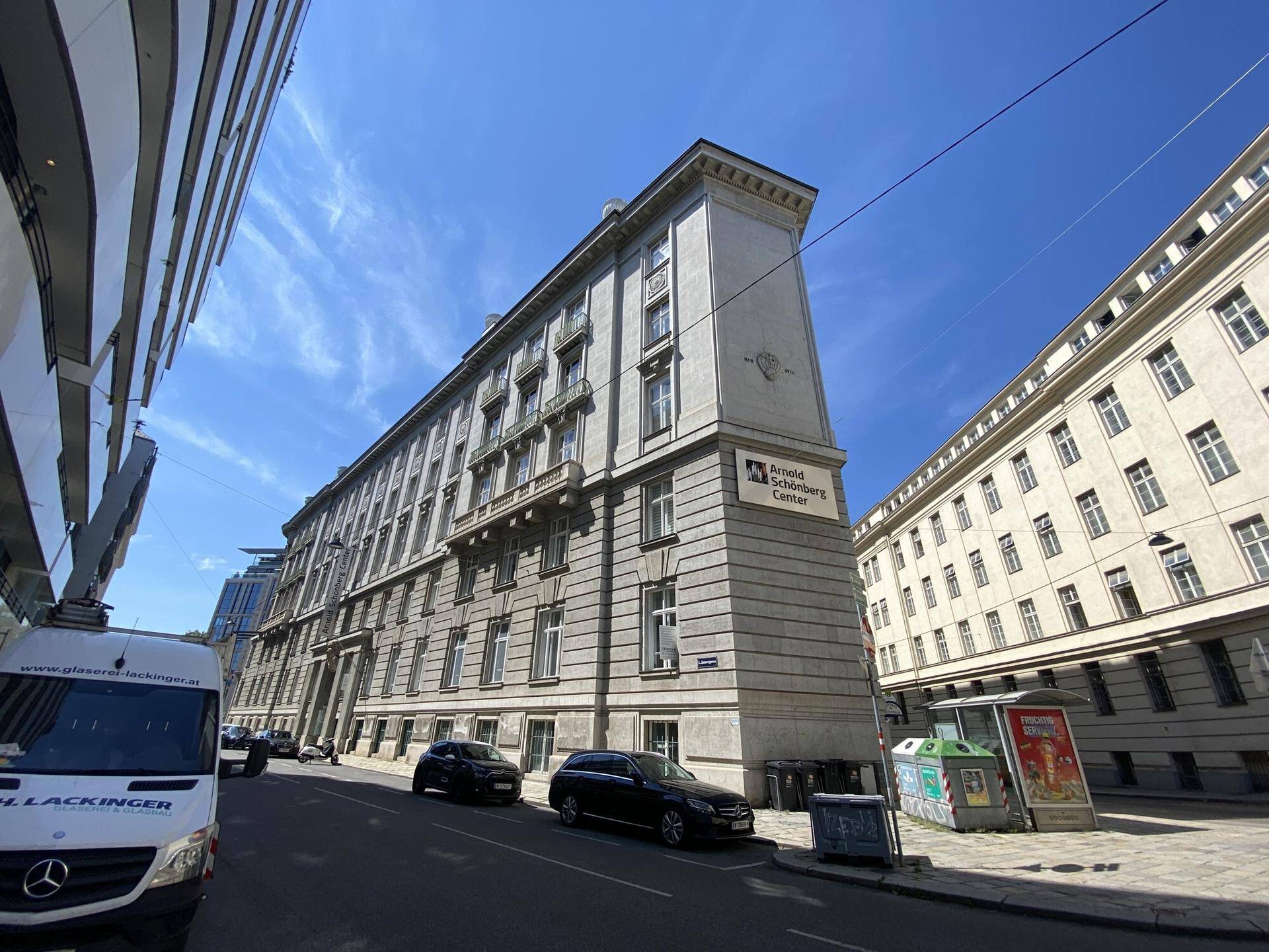 Klassisches Gebäude an einer belebten Straße unter blauem Himmel, mit parkenden Autos davor.