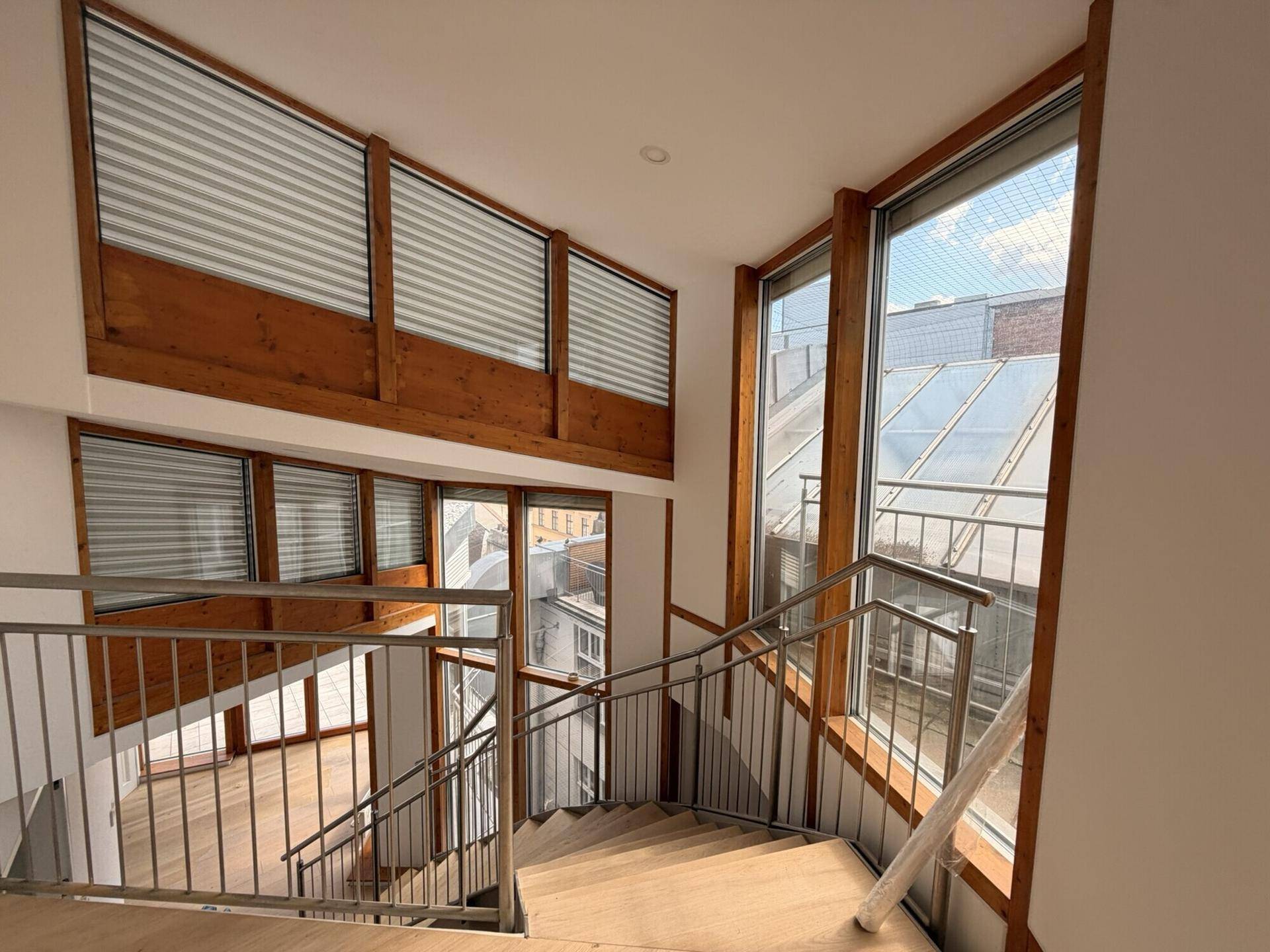 Blick von oben auf eine moderne Holztreppe mit Metallgeländer und großen Fenstern mit Jalousien.