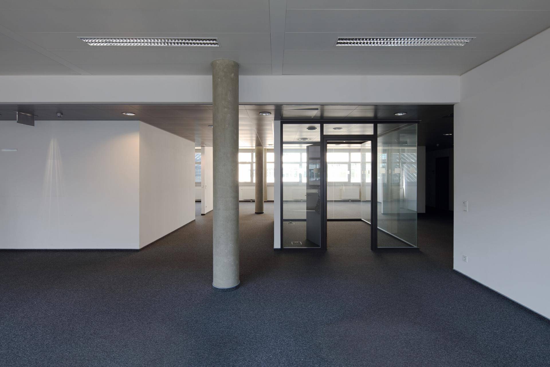 Leerer Büroraum mit Teppichboden, einer Betonsäule und Glaswänden, die verschiedene Bereiche abgrenzen.