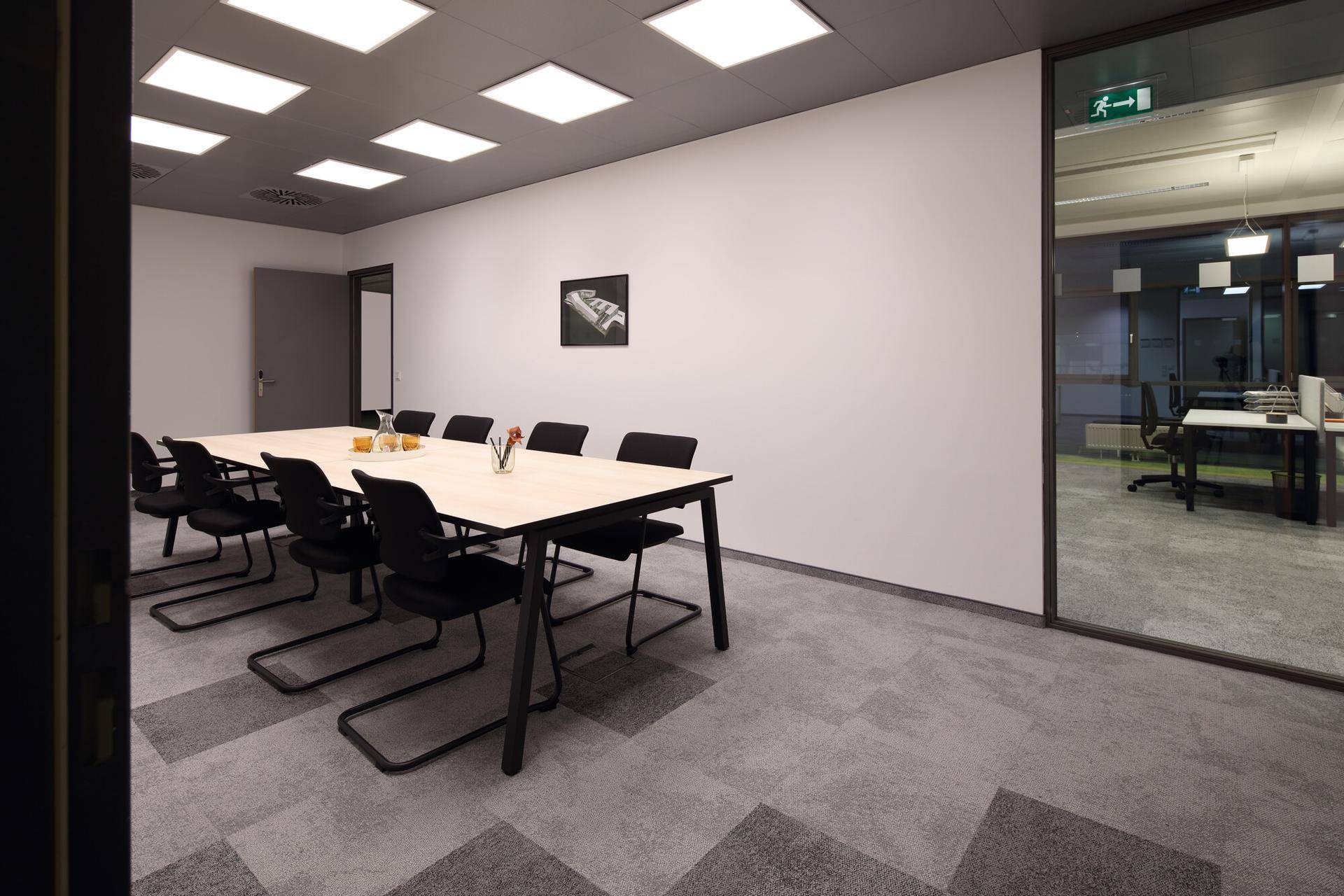 Moderner Konferenzraum mit großem Tisch, schwarzen Stühlen und Teppichboden, durch Glaswand getrennt.