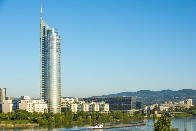 Modernes Hochhaus am Flussufer mit umliegenden Gebäuden und Bergen im Hintergrund.