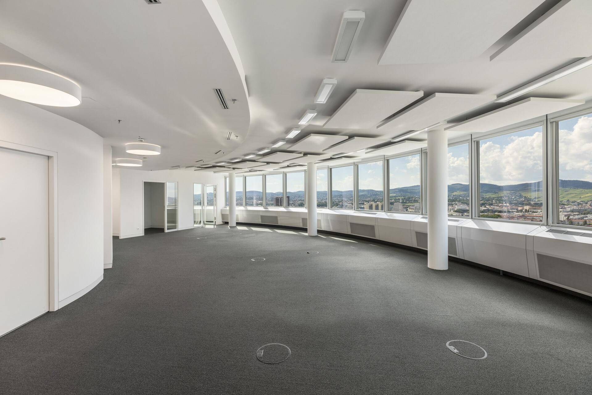Großes, leeres Büro mit Teppichboden, vielen Fenstern und Panoramablick auf die Stadt und Hügel.