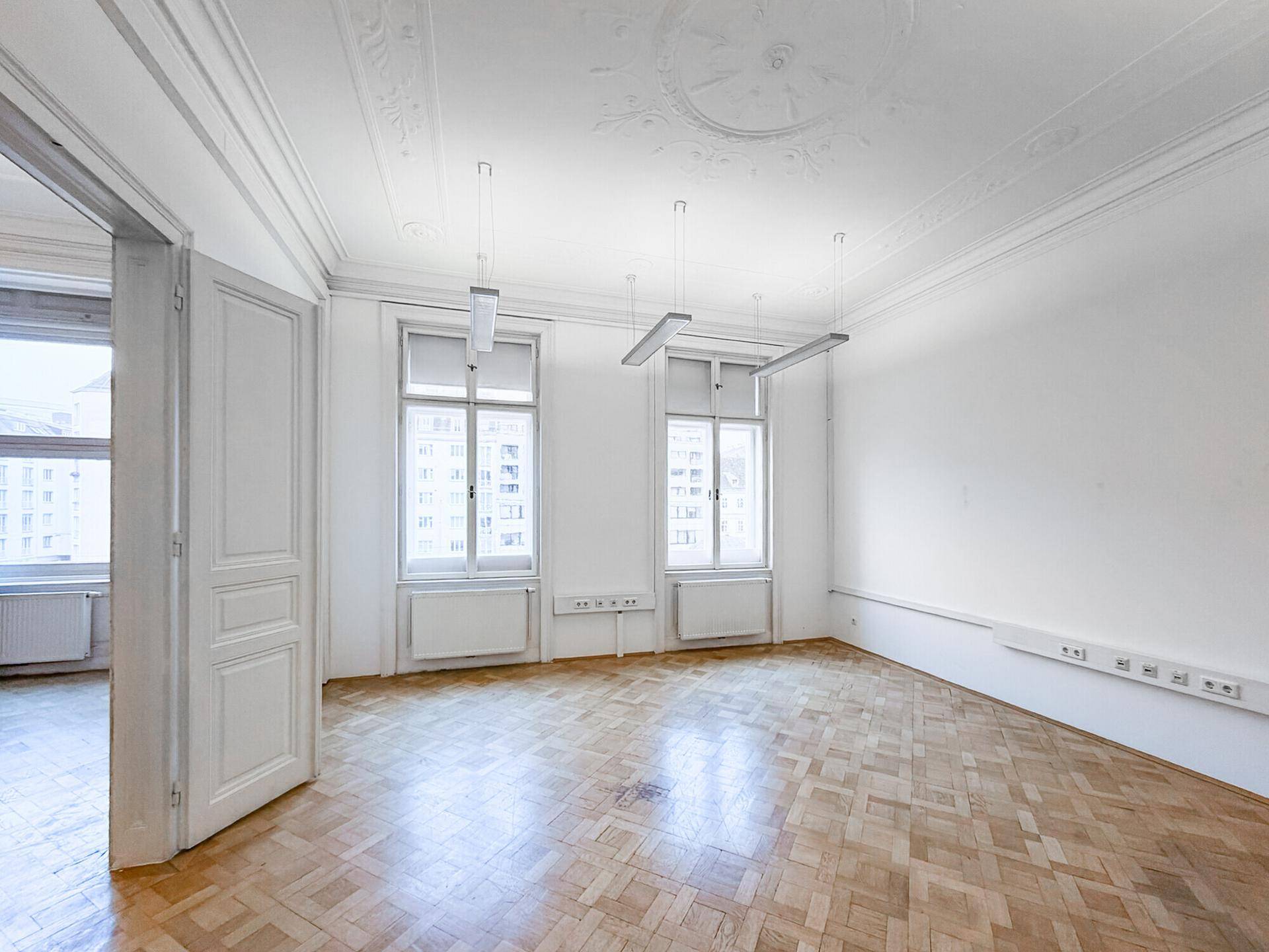 Leeres Büro mit Fischgrätparkett, hohen Fenstern und dekorativer Stuckverzierung an der Decke.