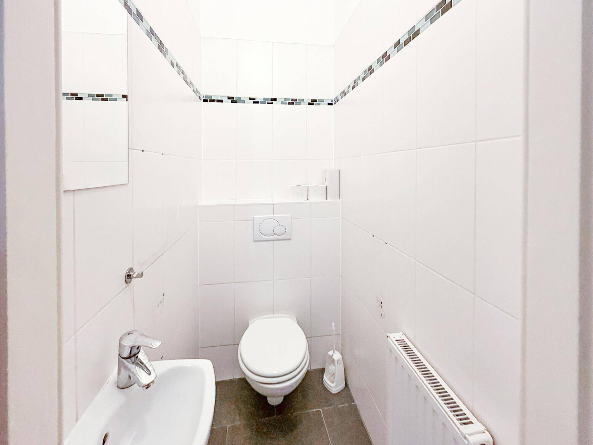 Separate Gästetoilette mit WC, kleinem Waschbecken und Heizkörper, weiß gefliest.