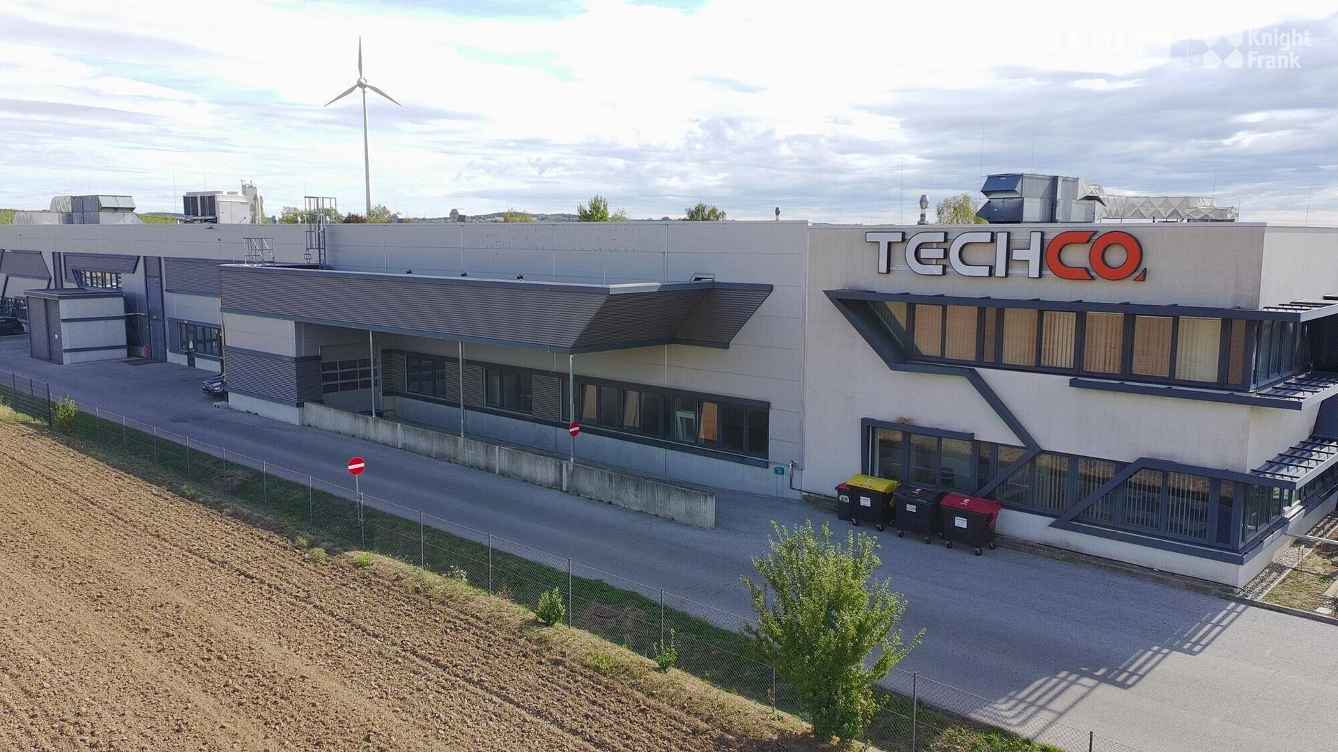 Moderne Gewerbeimmobilie mit Windrad im Hintergrund, Parkplätzen und weitem Blick über die Landschaft.