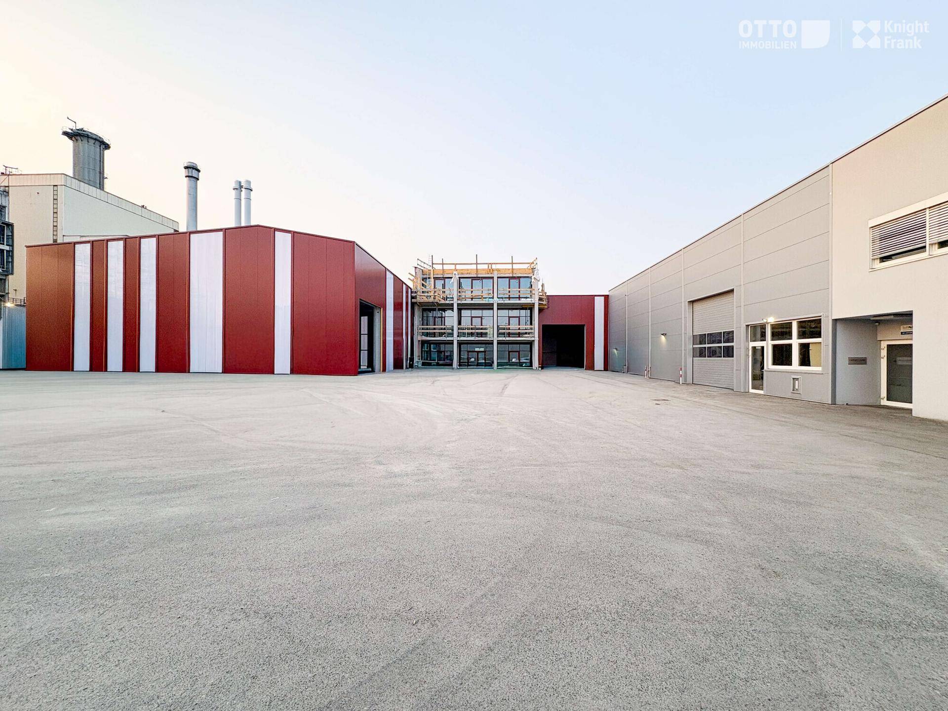 Außenansicht eines modernen Gewerbekomplexes mit roten und grauen Gebäuden sowie einer großen Freifläche.