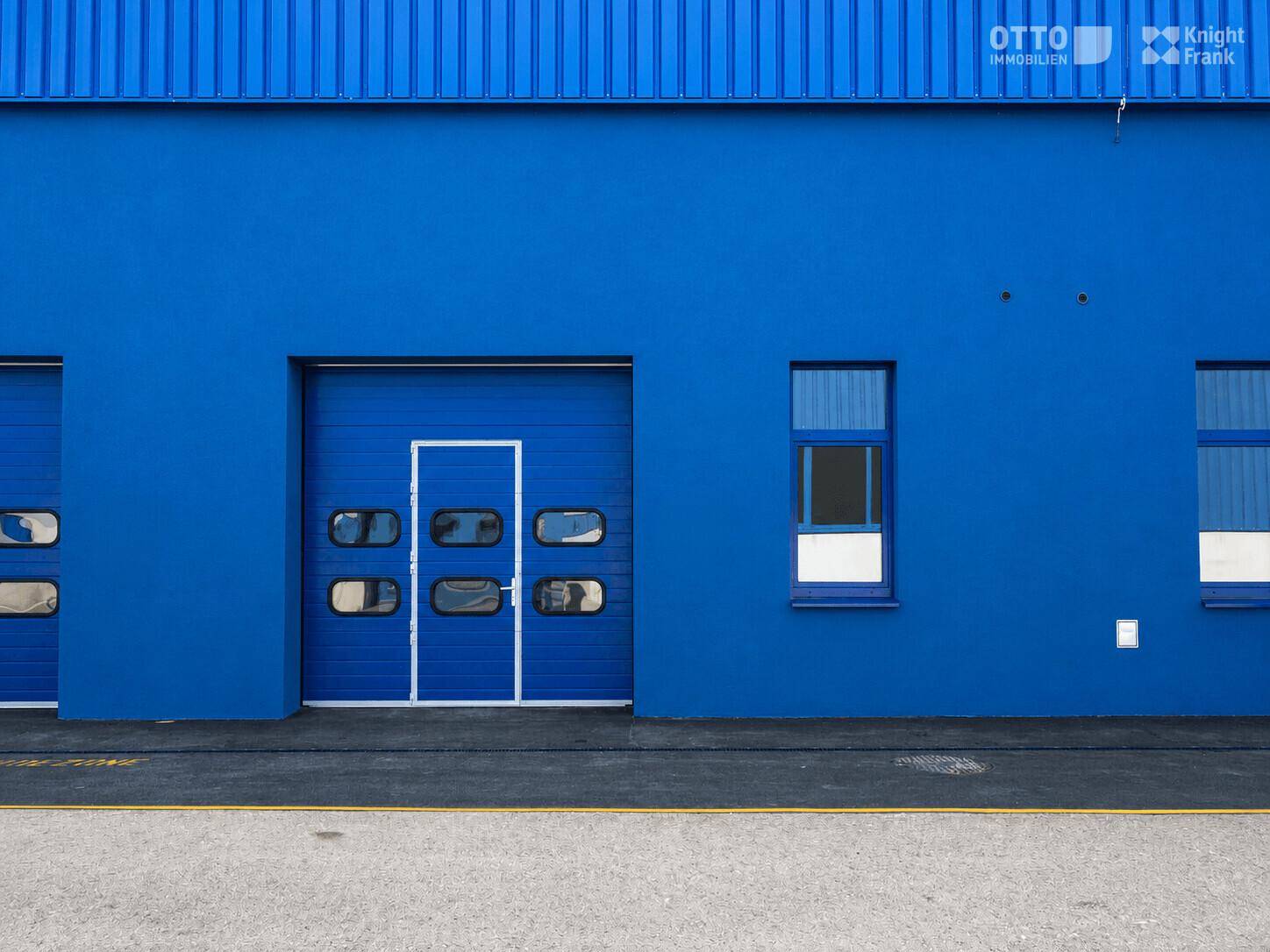 Blaue Fassade eines Industriegebäudes mit einem Rolltor und einem Fenster, das Tageslicht hereinlässt.