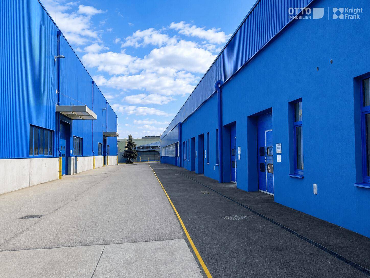 Weitläufiger Außenbereich zwischen zwei blauen Industriehallen unter einem bewölkten Himmel.