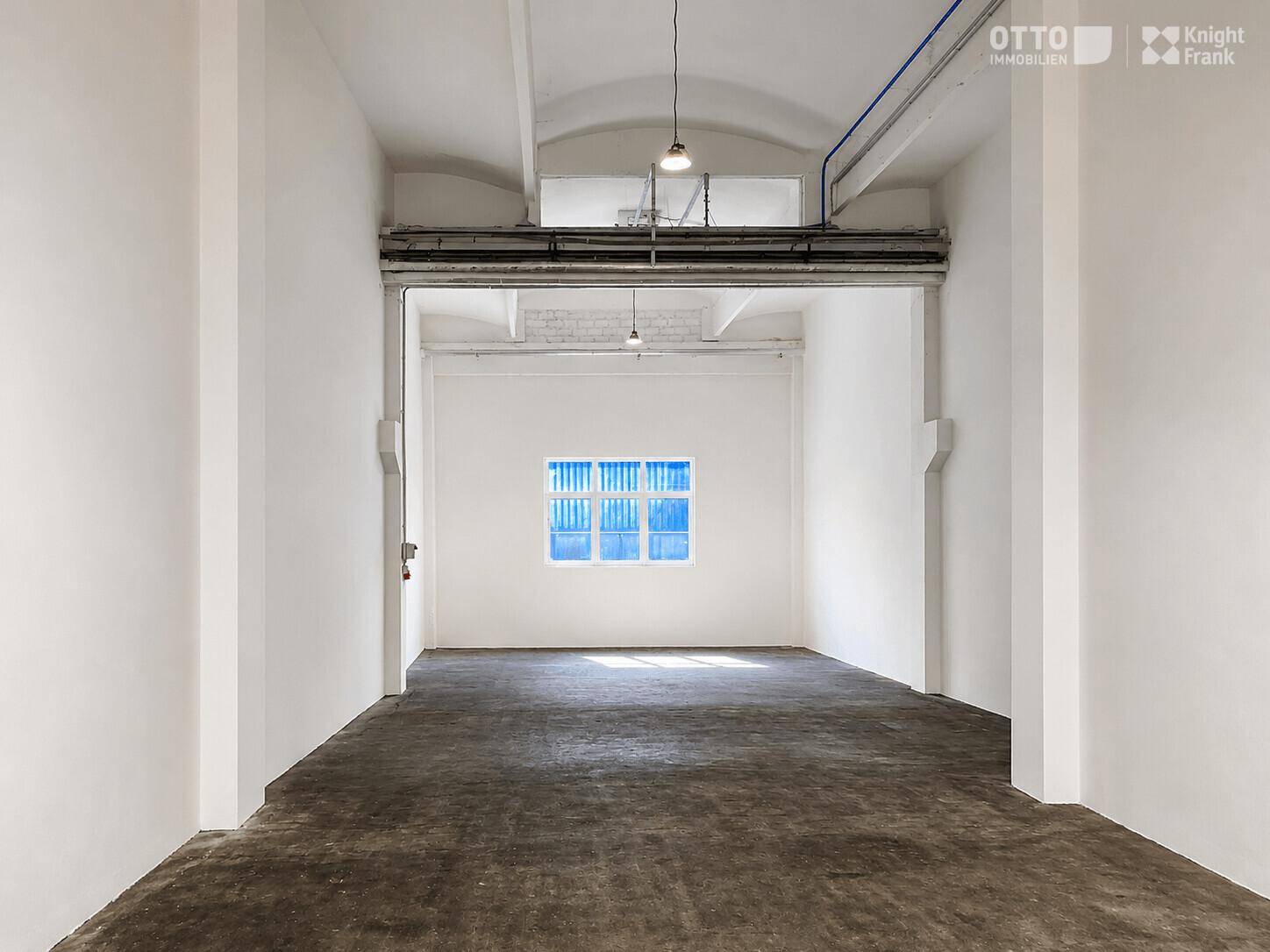 Geräumiger, leerer Lagerraum mit Betonboden und einem blauen Fenster, das Tageslicht hereinlässt.