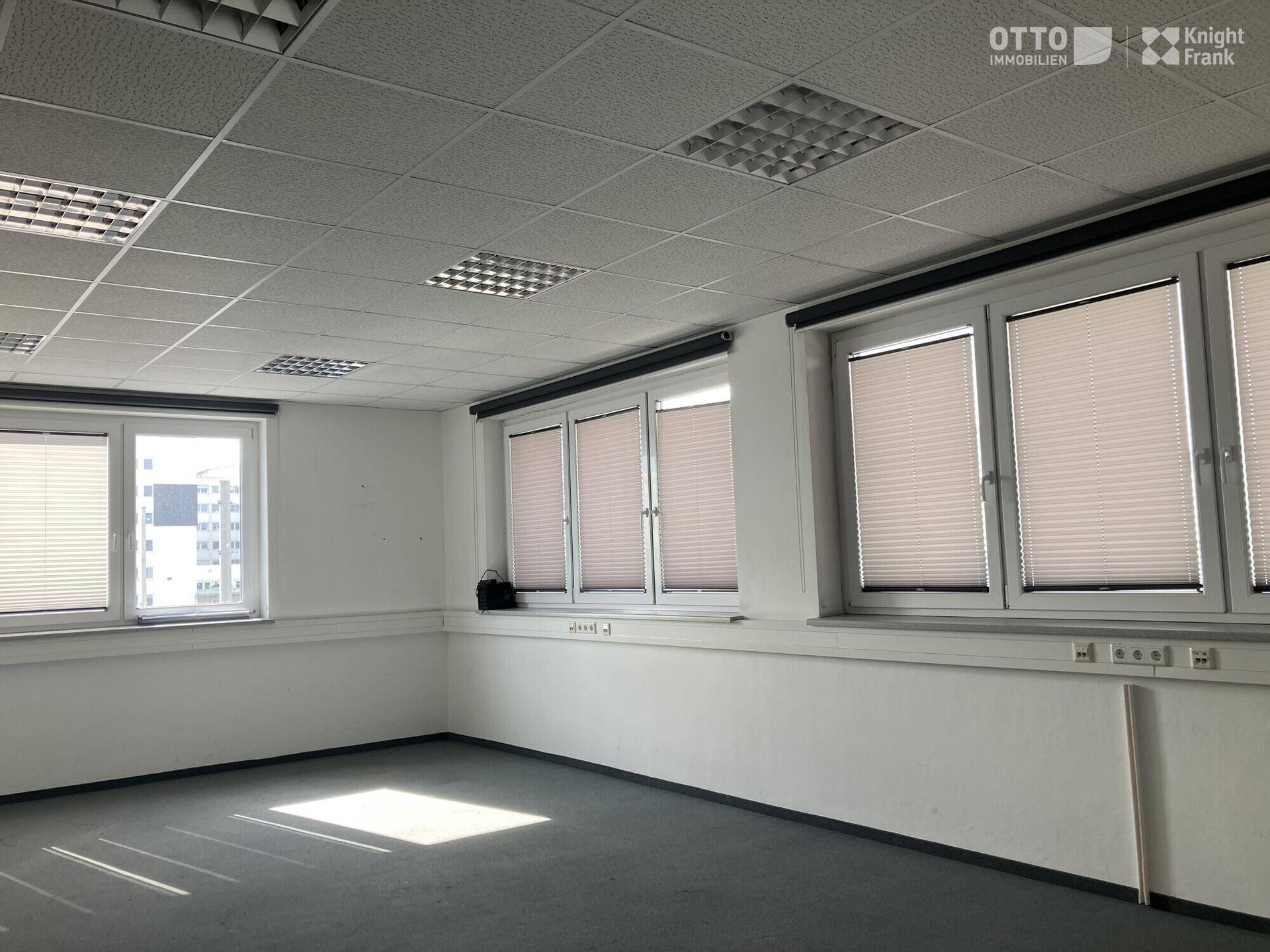 Büro mit Teppichboden, großen Fenstern mit Jalousien und einem weiten Blick auf die Umgebung.