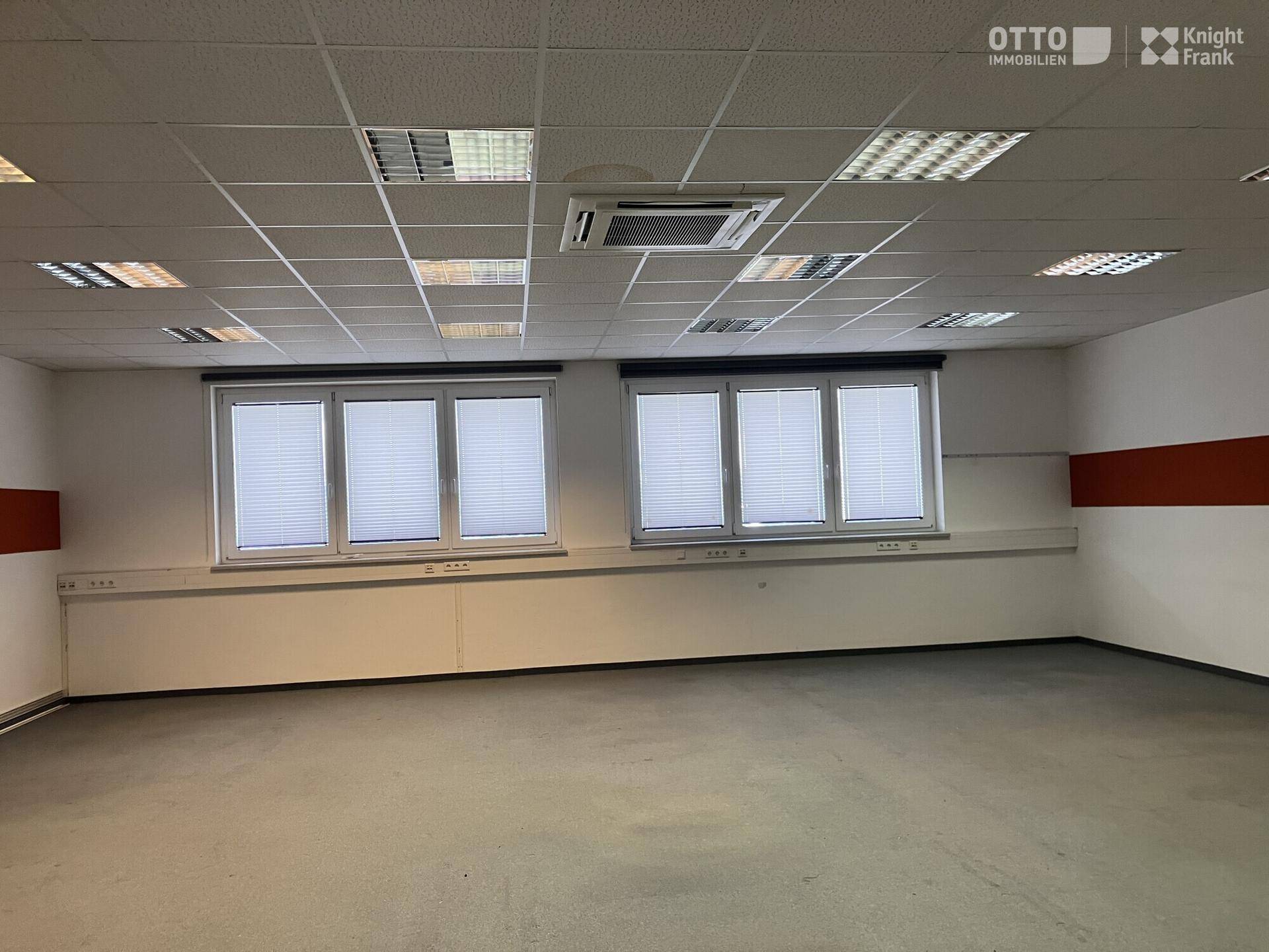 Helles Büro mit abgehängter Decke, zwei großen Fenstern mit Jalousien und einer Klimaanlage.