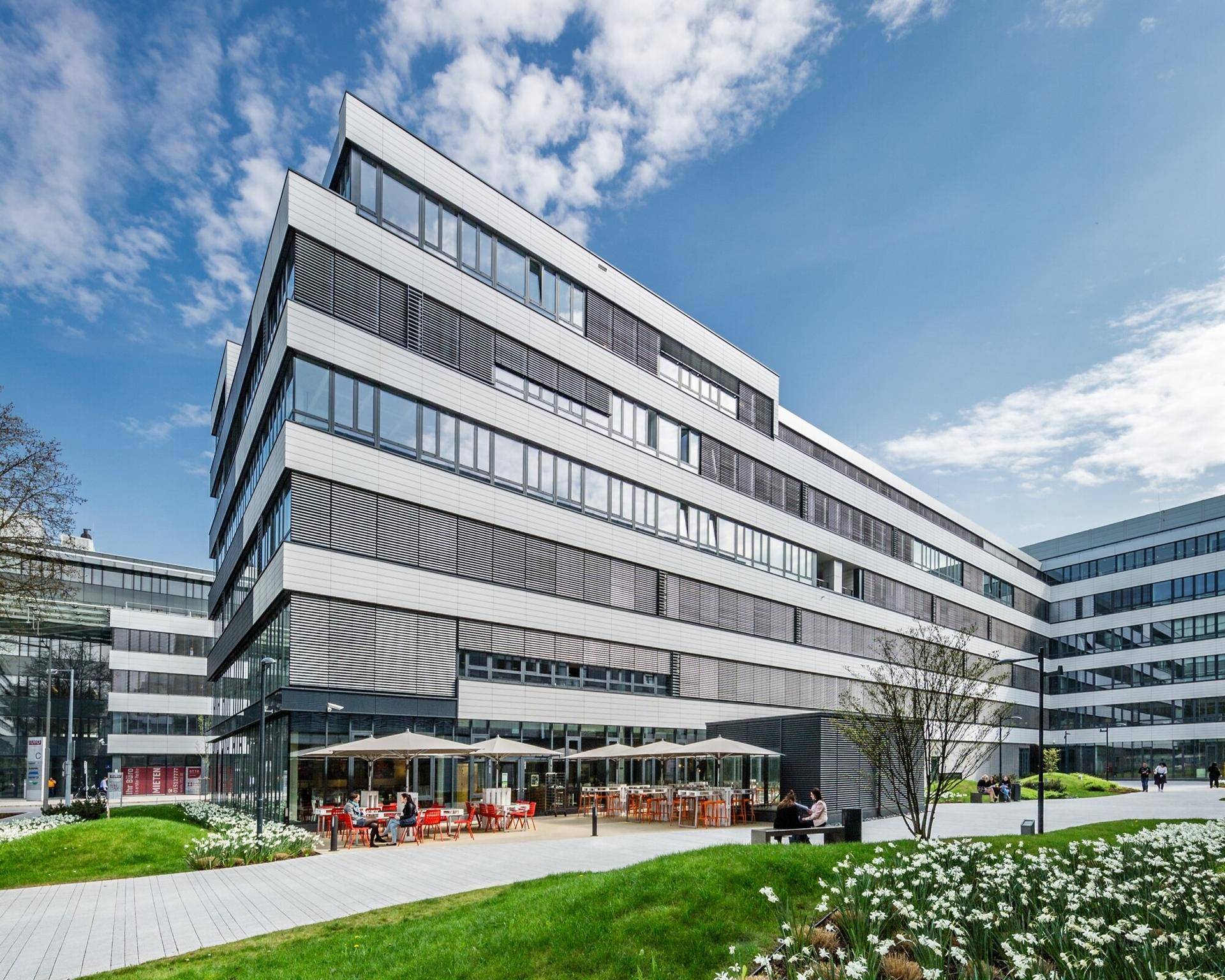 Außenansicht eines modernen Bürogebäudes mit einer belebten Café-Terrasse im Erdgeschoss.