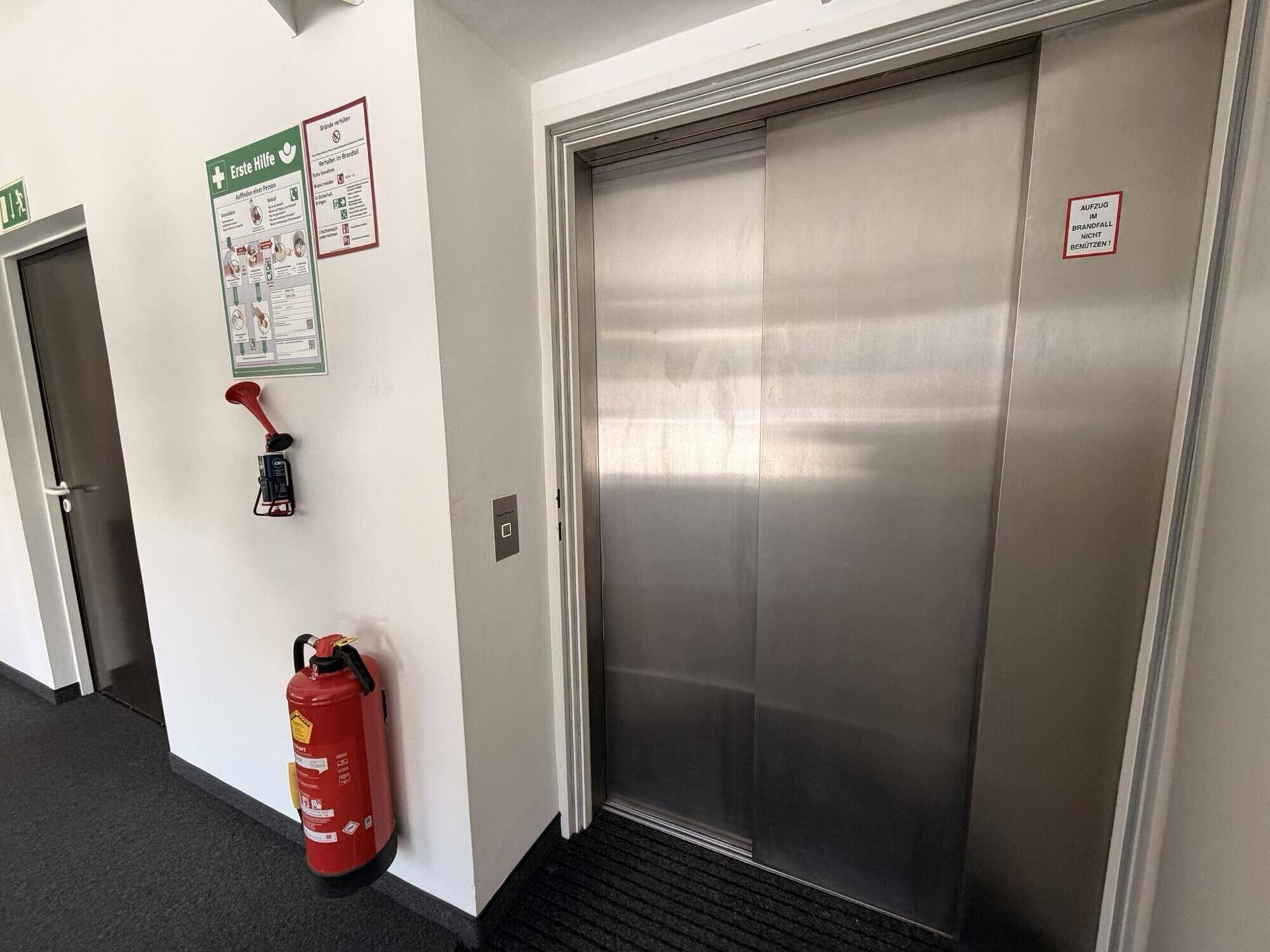 Flur mit einem modernen Aufzug, einem Feuerlöscher und Sicherheitshinweisen an der Wand.