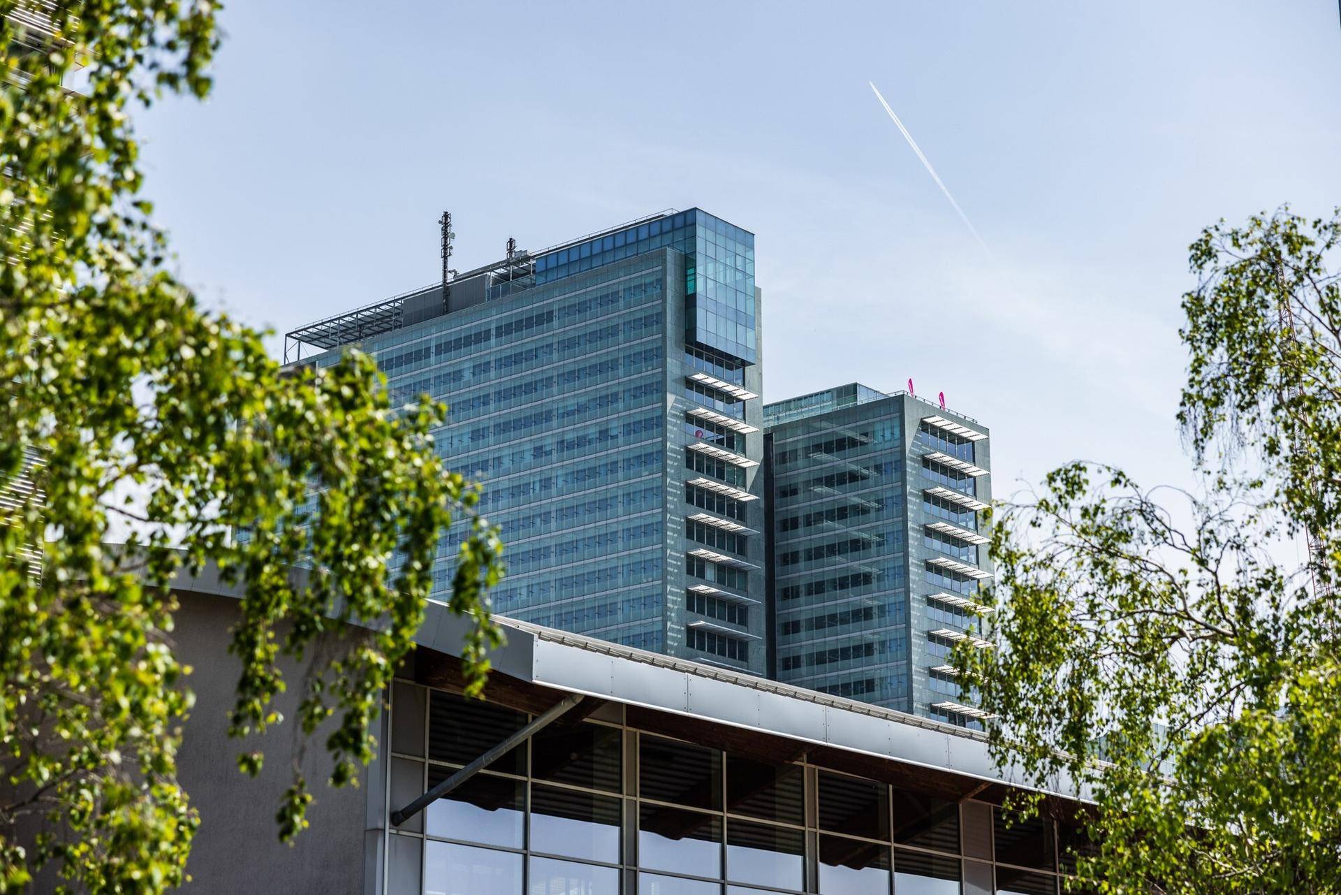 Moderner Wolkenkratzer mit Glasfassade und mehreren Etagen, umgeben von Bäumen unter blauem Himmel.