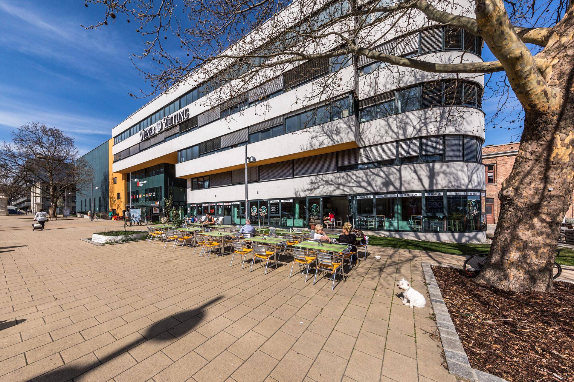 Außenansicht eines modernen Bürogebäudes mit belebter Café-Terrasse und Sitzgelegenheiten im Freien.