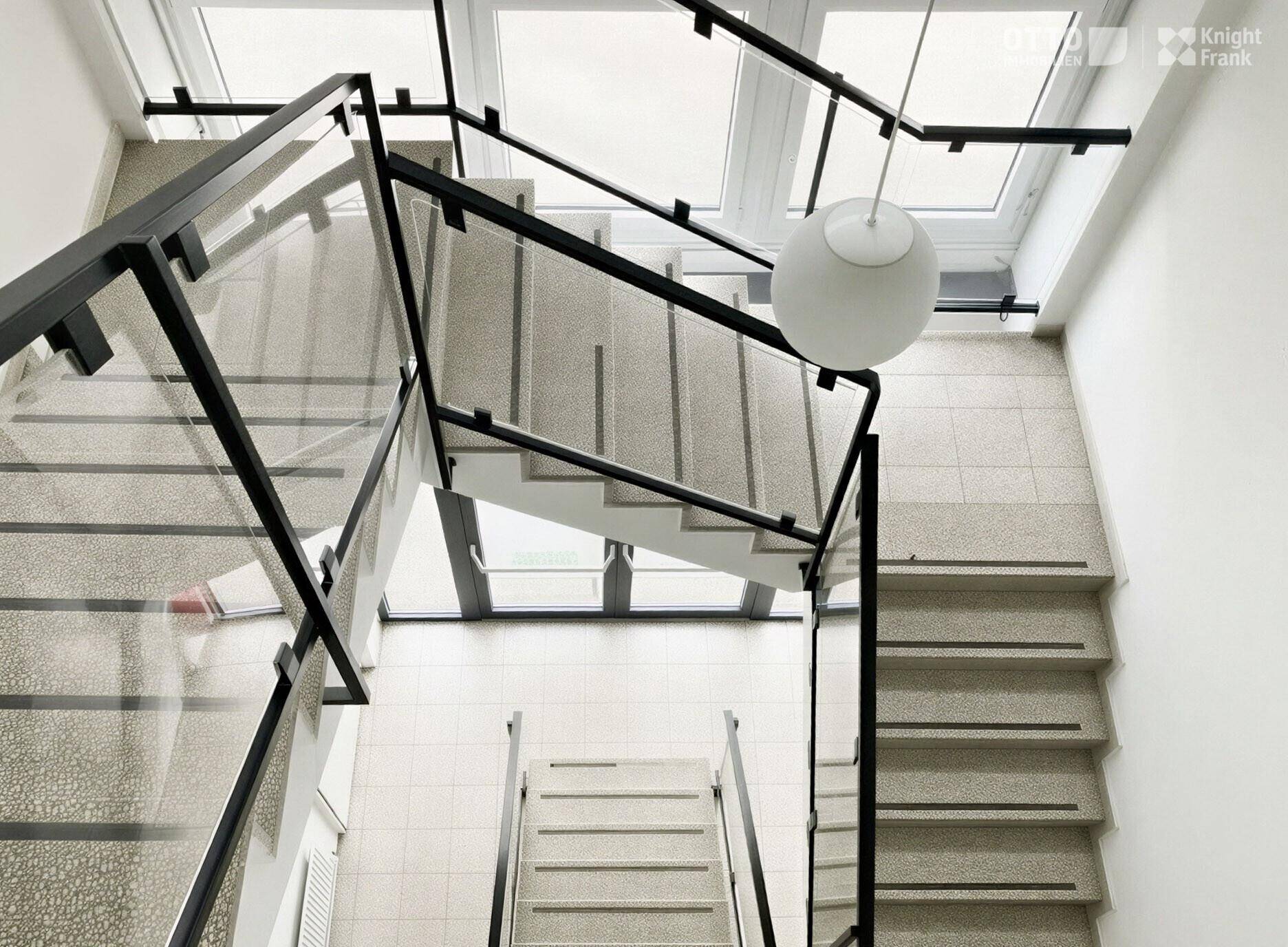 Modernes Treppenhaus mit Glasgeländer, hellen Stufen und natürlichem Lichteinfall von oben.