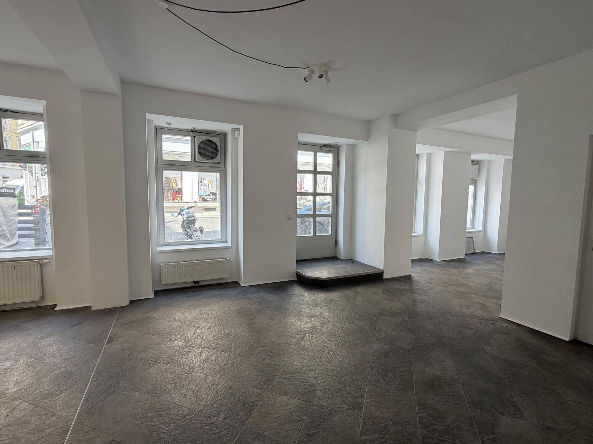 Heller, leerer Raum mit großen Fenstern zur Straße, dunklem Fliesenboden und Klimaanlage.