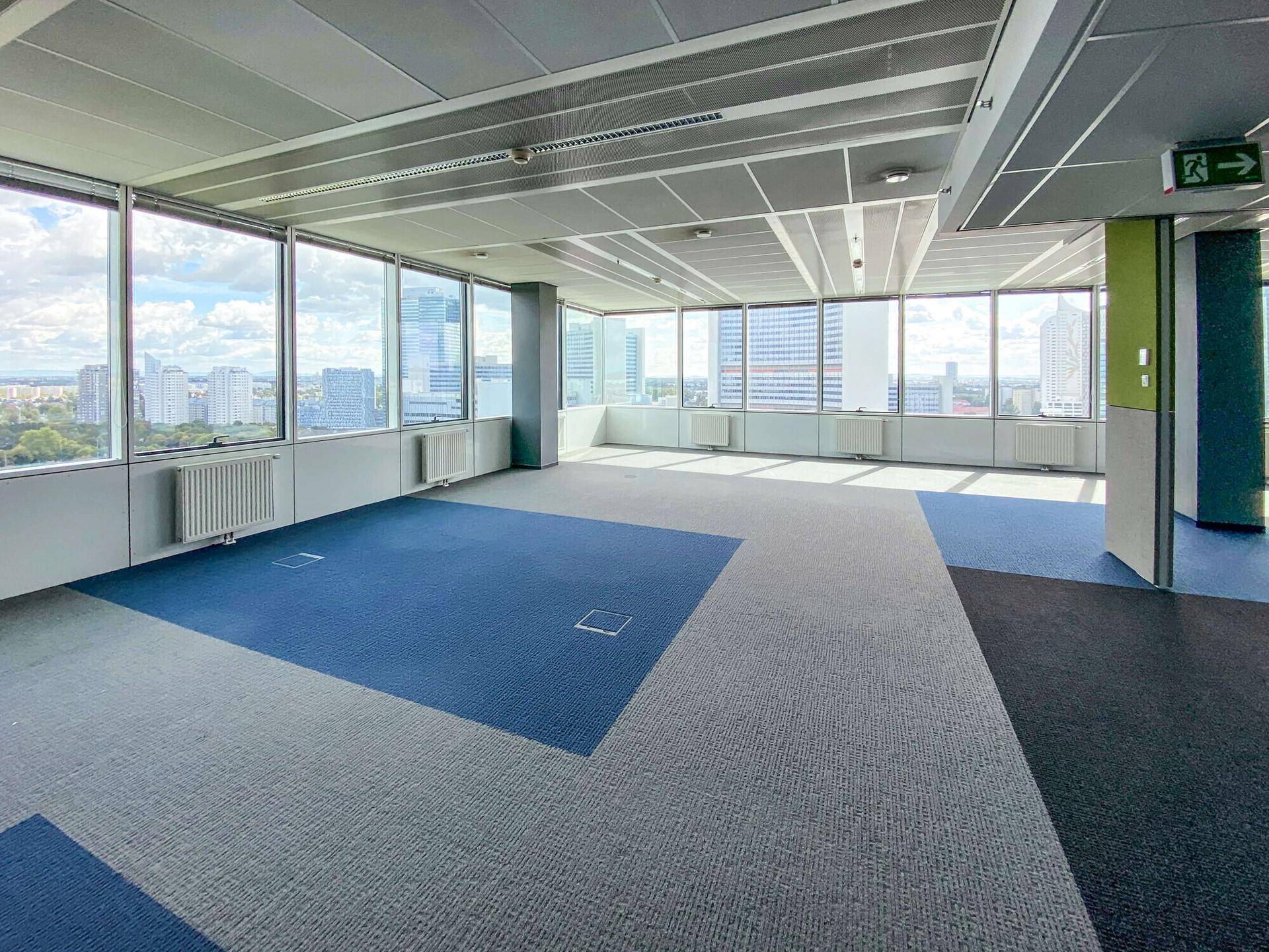 Helles Büro mit großen Fenstern, blau-grauem Teppichboden und Blick auf die umliegende Stadt.