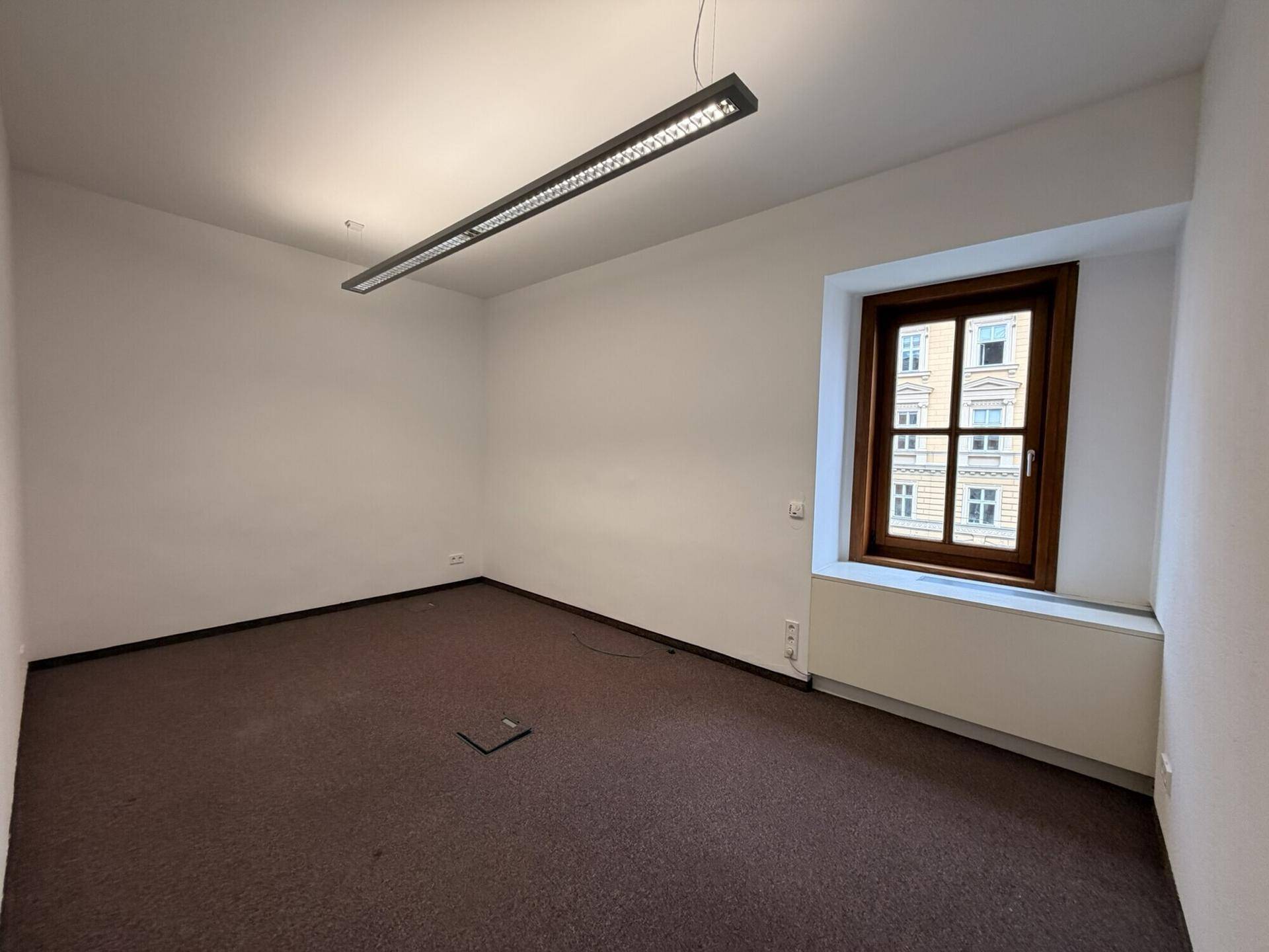 Kleiner Büroraum mit braunem Teppichboden und einem Fenster, das Tageslicht spendet.
