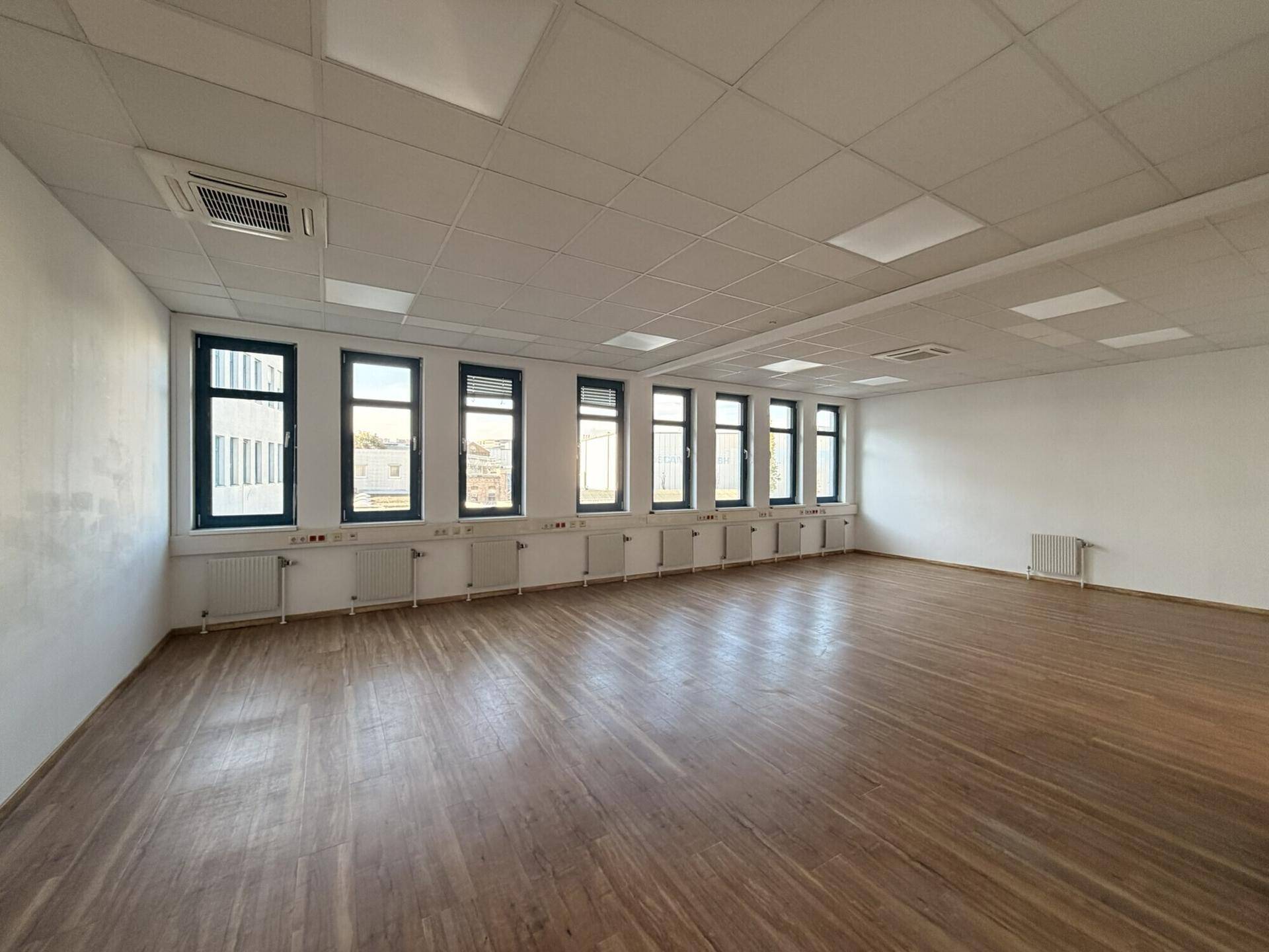 Geräumiger, leerer Büroraum mit Holzboden, vielen Fenstern und abgehängter Decke.