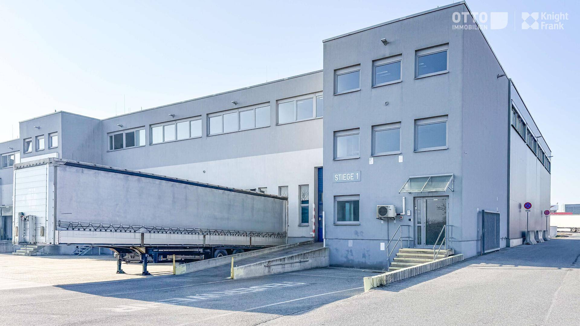 Modernes Gewerbegebäude mit Laderampen, einem Lastwagenanhänger und einem Eingangsbereich mit Treppe.