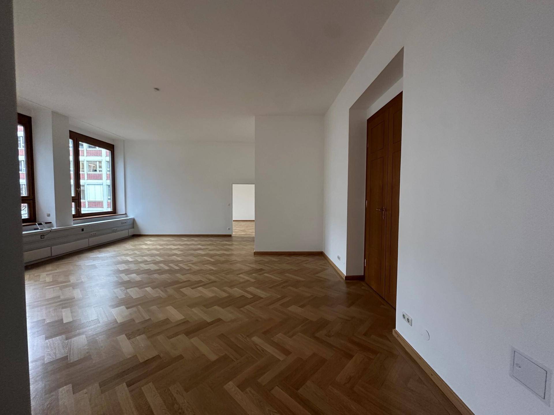 Großer, heller Raum mit Fischgrätparkett und hohen Fenstern, ideal für Wohn- oder Bürozwecke.