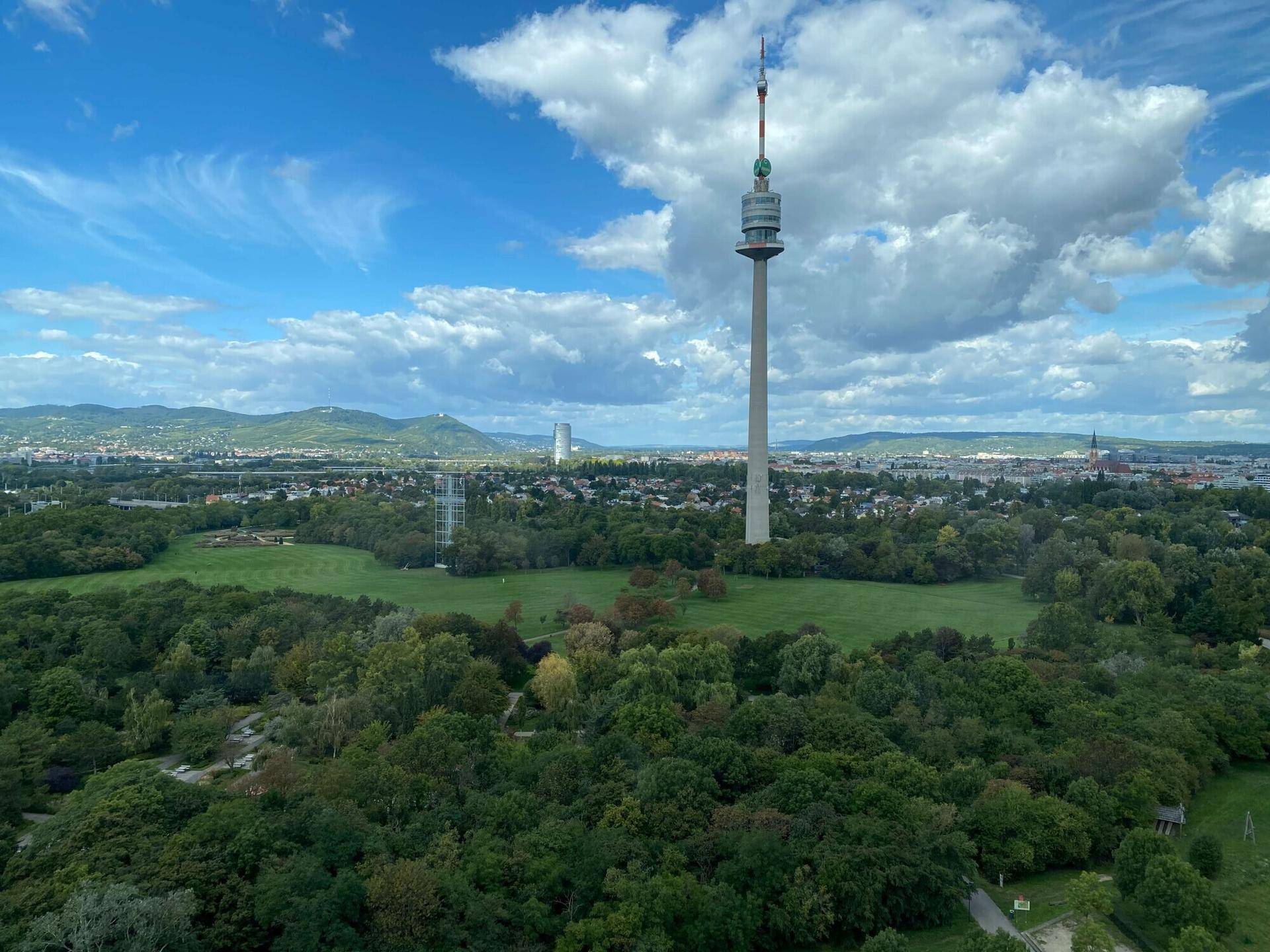 Panoramablick auf den Donauturm, weite Grünflächen und die Stadtlandschaft unter einem blauen Himmel.