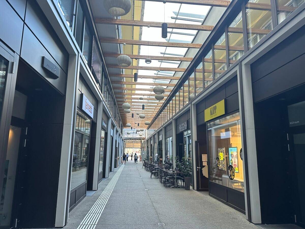 Überdachte Ladenpassage mit Geschäften, Sitzgelegenheiten und einer modernen Glasdachkonstruktion.