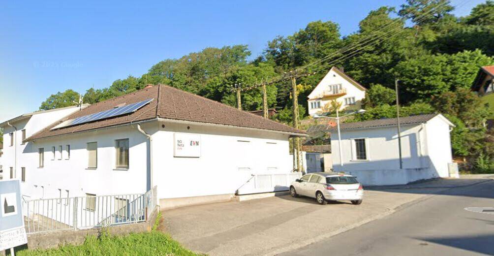 Weißes Gebäude mit braunem Dach und Photovoltaikanlage, umgeben von grüner Landschaft.