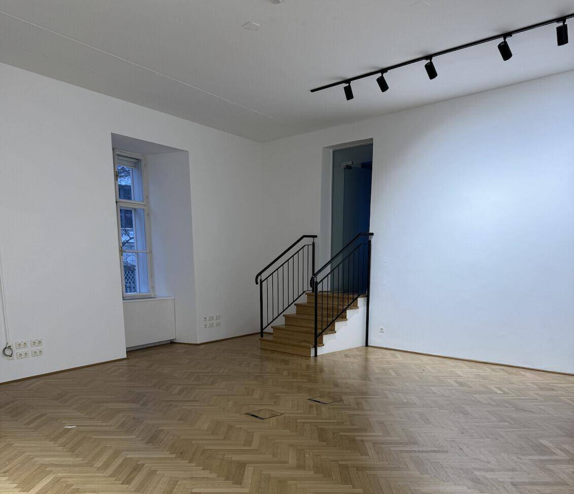 Leerer Flur mit Fischgrätparkett, einem Fenster und einem Treppenaufgang, ideal für eine vielseitige Nutzung.