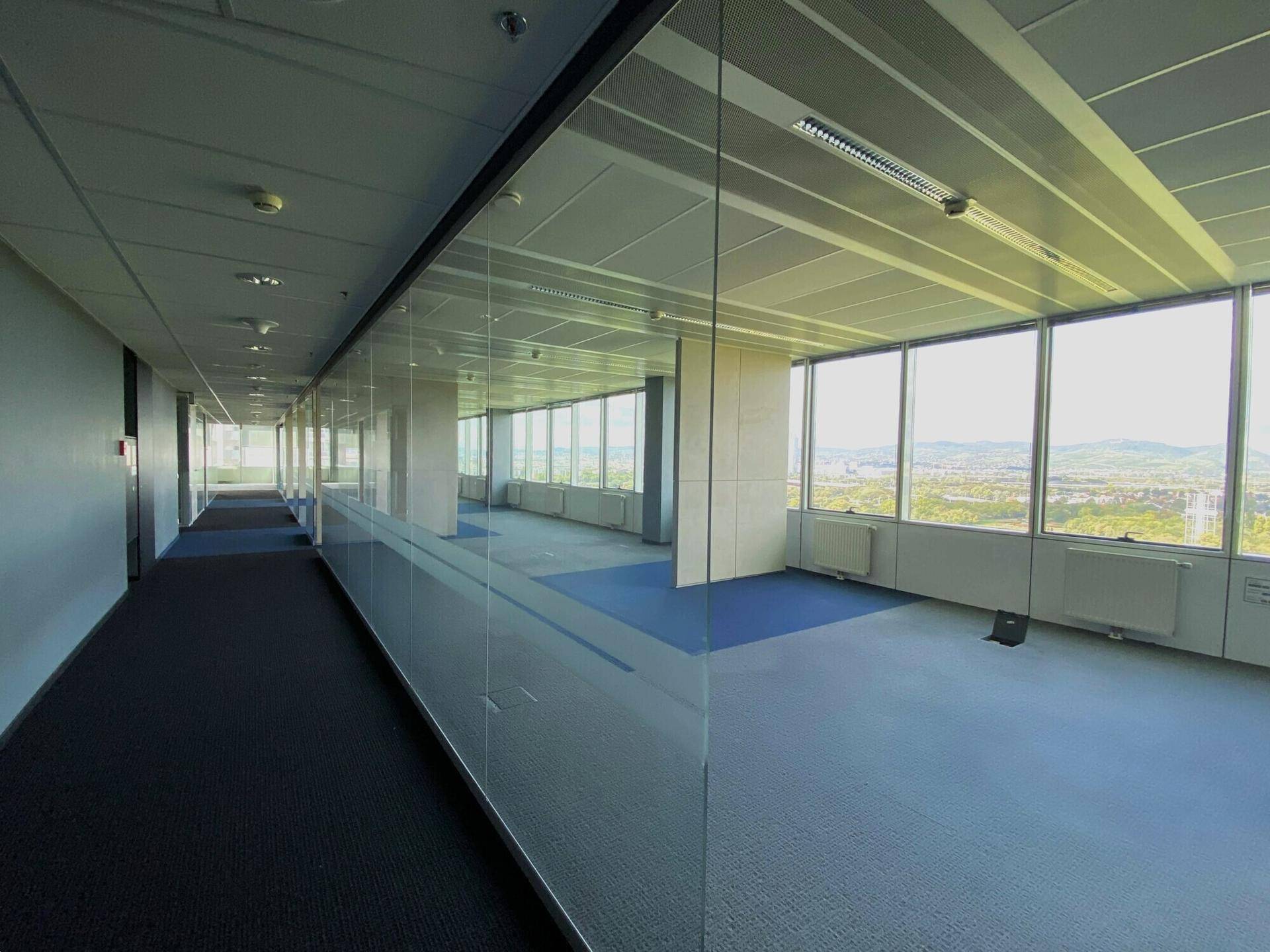 Langer Bürogang mit Glaswänden, die den Blick in offene Büroräume und auf die Stadtlandschaft freigeben.