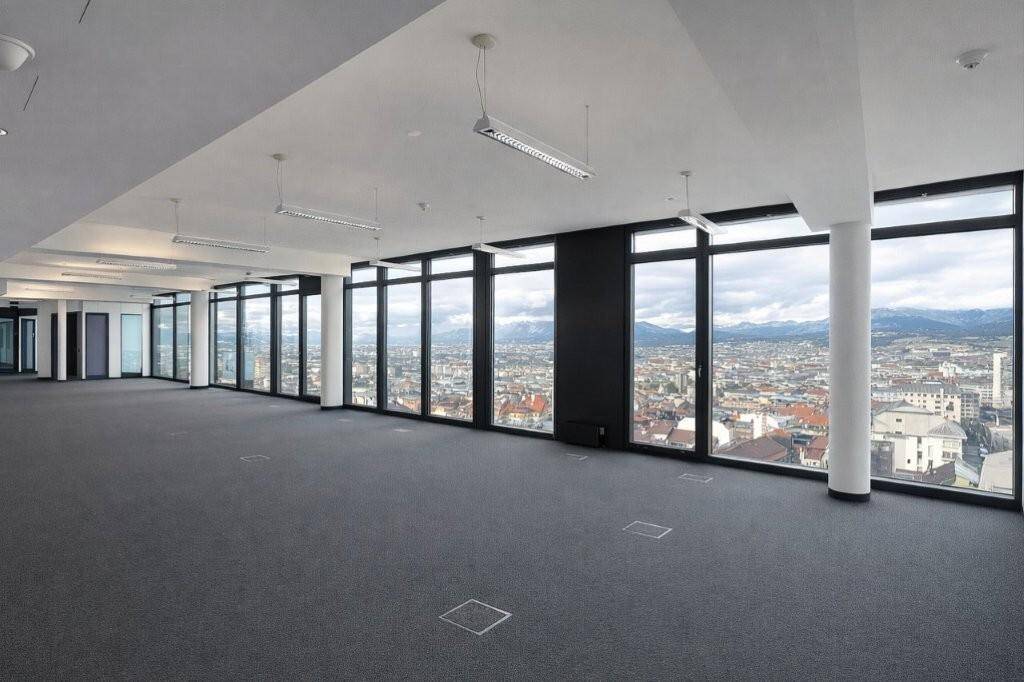 Geräumiger, leerer Büroraum mit Teppichboden und raumhohen Fenstern, die einen weiten Stadtblick bieten.