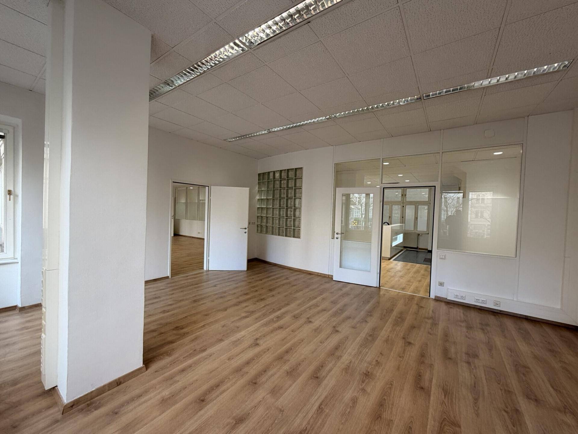 Großes Büro mit Laminatboden, Glasbausteinwand und Zugang zu weiteren, hellen Räumen.